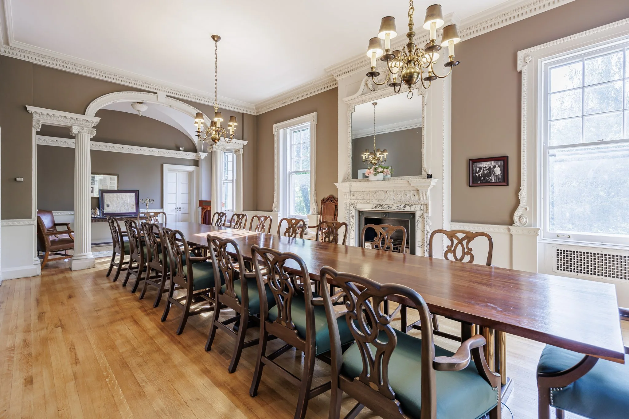 Elegant dining room with a long wooden table surrounded by wooden chairs, a marble fireplace, large windows, and ornate molding.