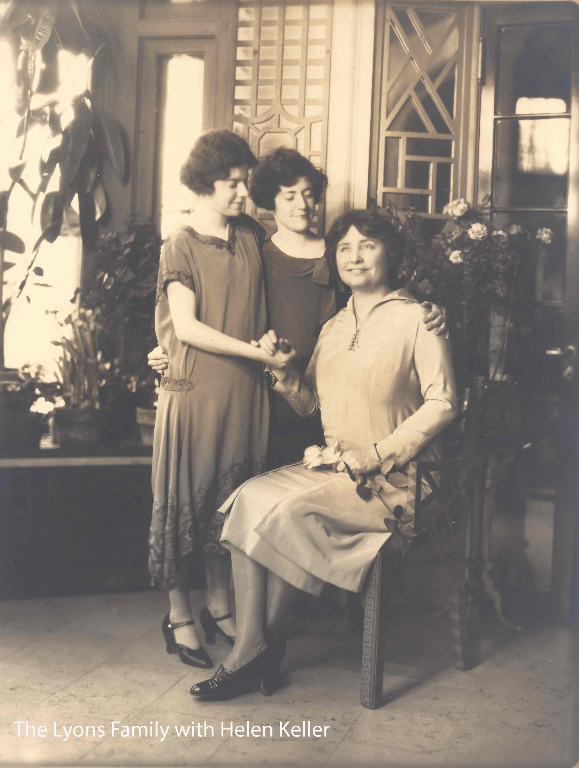 Black and white photo of three women, with one seated and two standing behind her, in an indoor setting with decorative wooden panels and plants. The seated woman is wearing a light-colored dress and dark shoes, holding a flower, and smiling at the camera. The standing women are dressed in dark dresses and are looking at the seated woman, with the woman in the middle holding her hand. The photo has the text "The Lyons Family with Helen Keller."