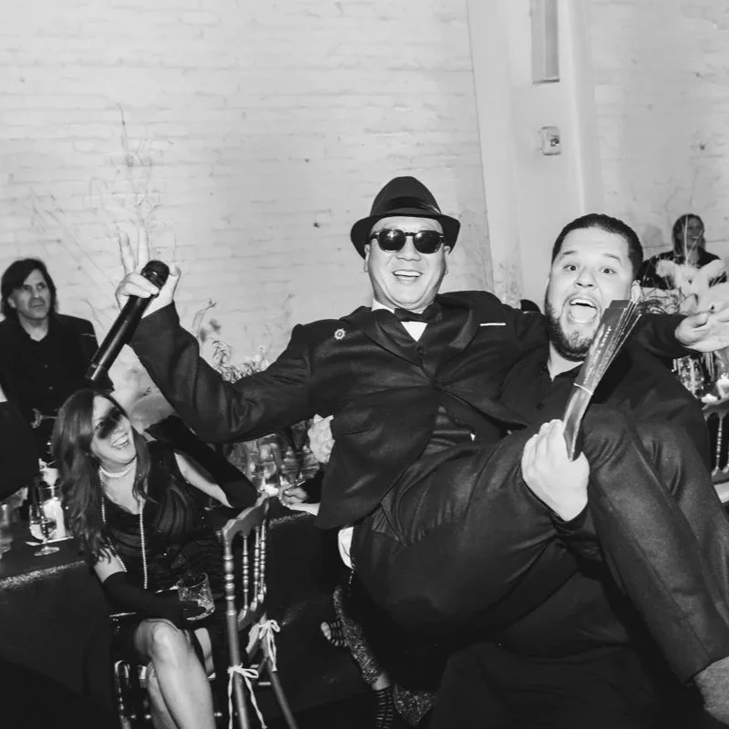 Two men celebrating at a party, one wearing sunglasses and a hat, holding a microphone, and the other holding a fan, with guests seated at tables in the background.