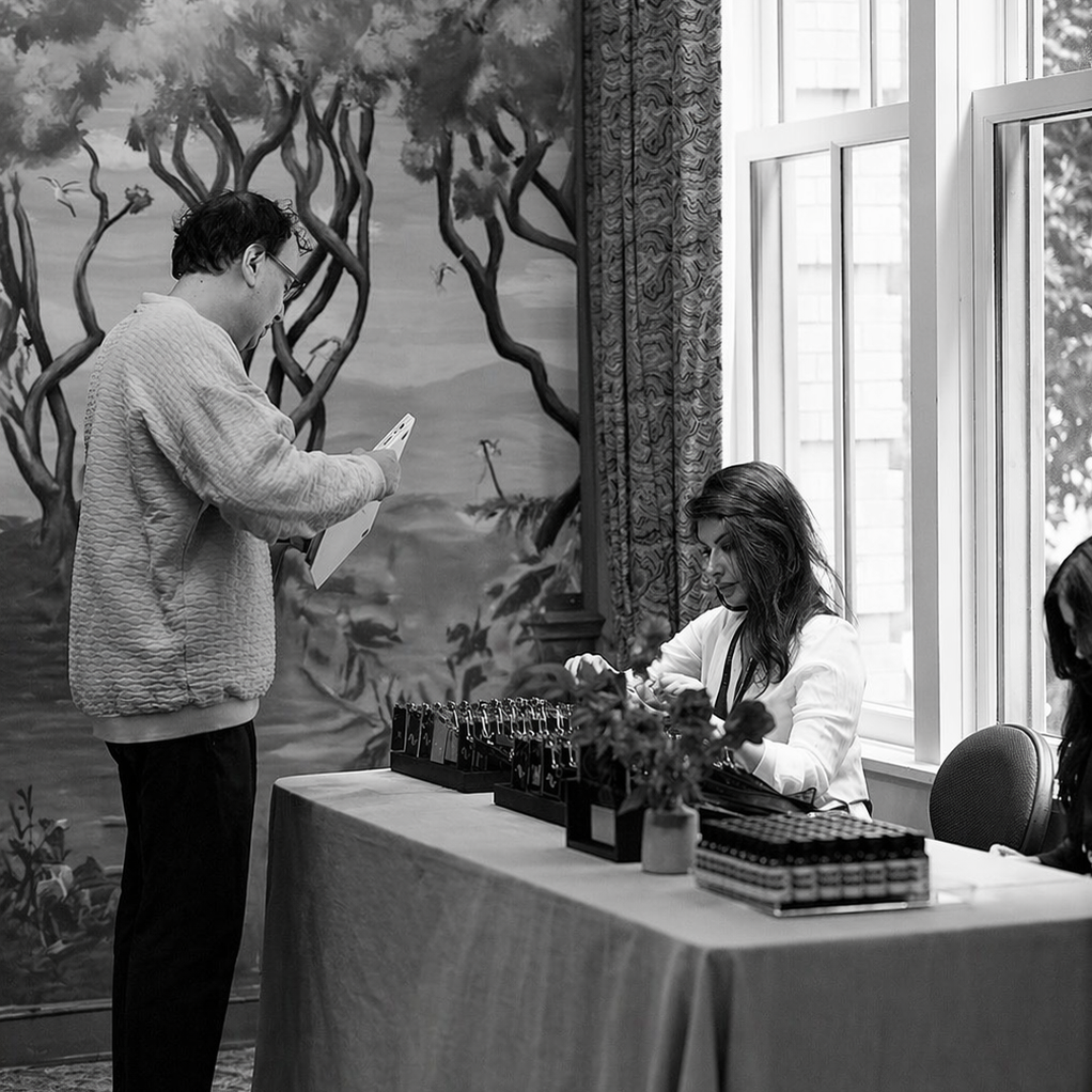 A man stands looking at a tablet at a table with various items, while a woman sits behind the table working with scratch-off maps or tickets next to a large window with curtains, in a room with a painted mural of trees.