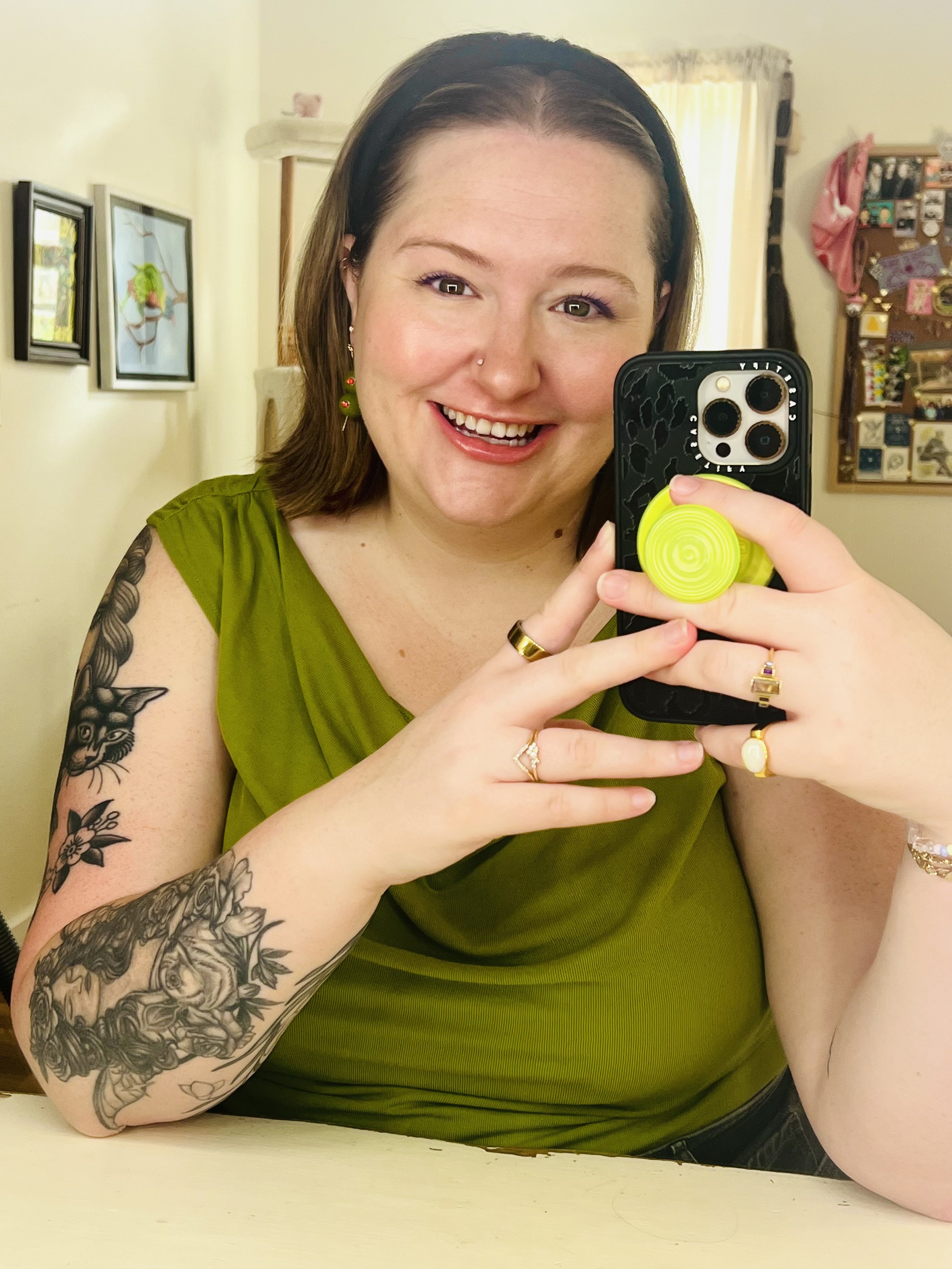 A woman with shoulder-length brown hair wearing a black headband and a green top, taking a mirror selfie. She has tattoos of flowers and a cat on her left arm, jewelry including earrings, rings, and a bracelet, and a nose piercing. She is smiling, showing her teeth, and holding a phone with a yellow pop socket. The background shows a brightly lit room with artwork on the wall and a corkboard with various items and a pink item hanging on it.