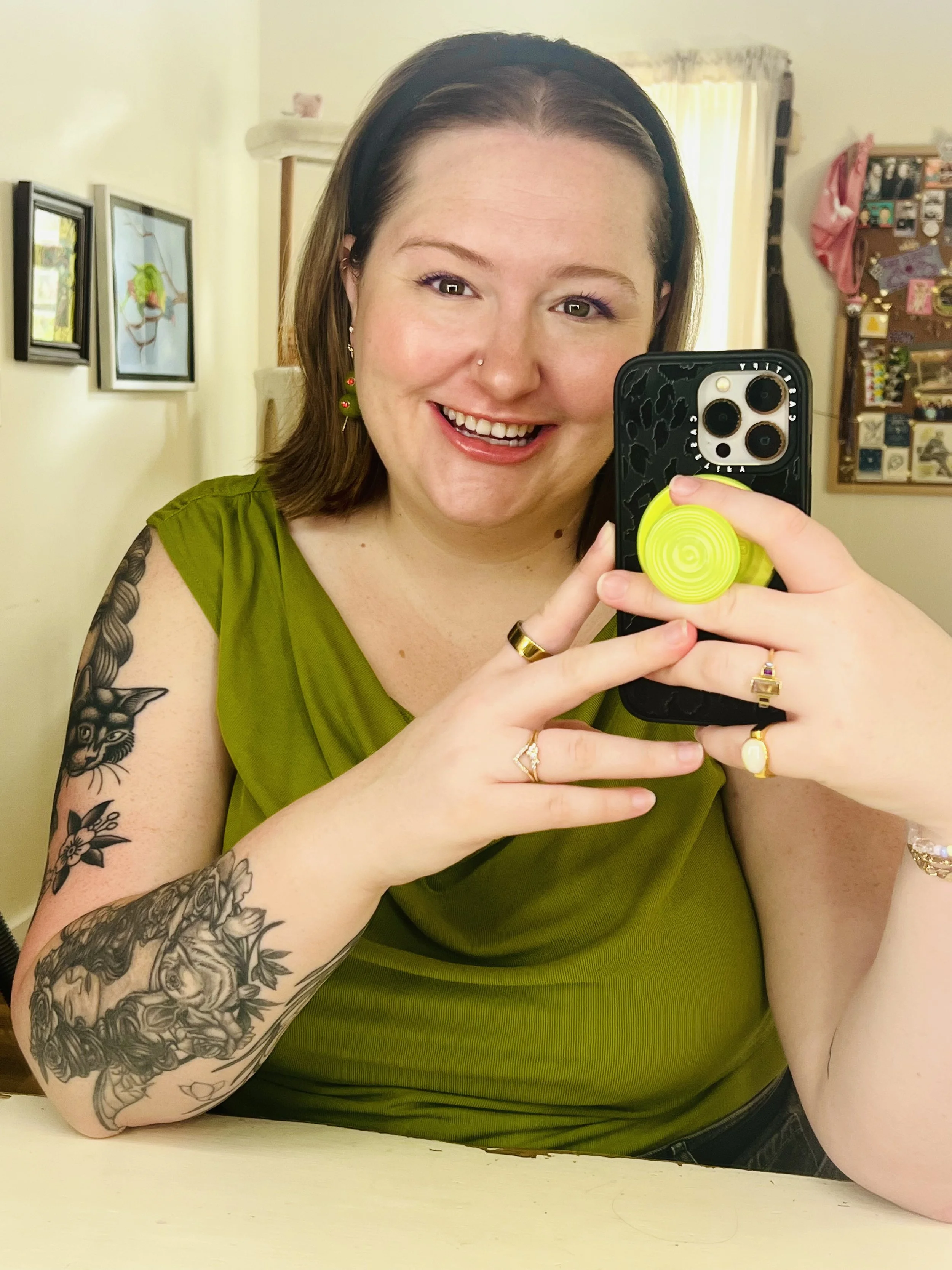 A woman smiling and taking a selfie with a smartphone. She has shoulder-length hair, wears a black headband, a green sleeveless top, and has tattoos on her left arm. She is wearing earrings, rings, and a bracelet. In the background, there are framed pictures on the wall and a corkboard with various items pinned on it.