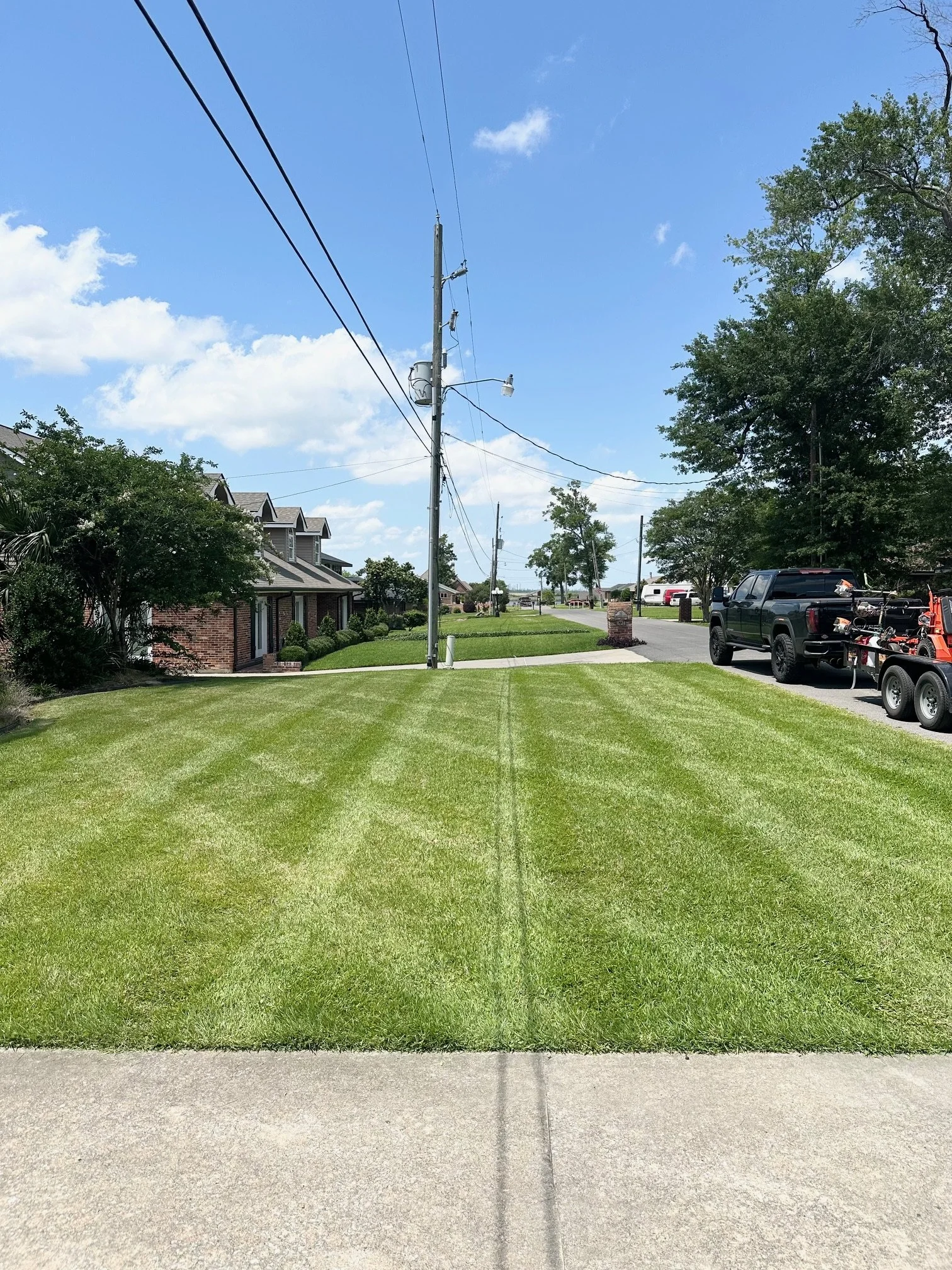 Bright green, freshly mowed lawn with tire marks, alongside a paved sidewalk and residential houses on the left and parked vehicles on the right. Overhead power lines extend into the distance under a partly cloudy blue sky.