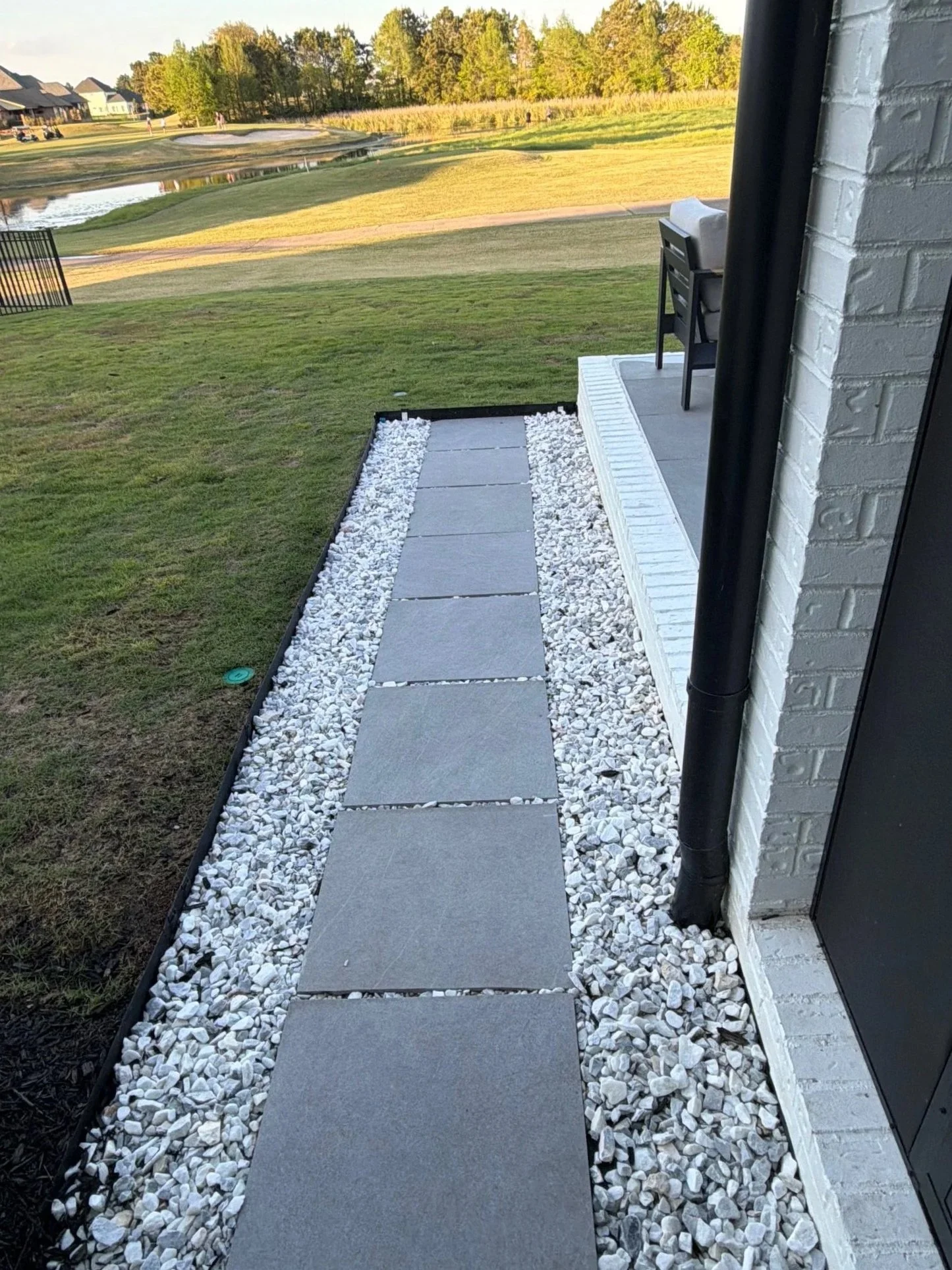 Concrete stepping stones on a narrow pathway with white gravel on either side, next to a white brick wall and a black drainpipe, leading to a patio overlooking a golf course and a pond.