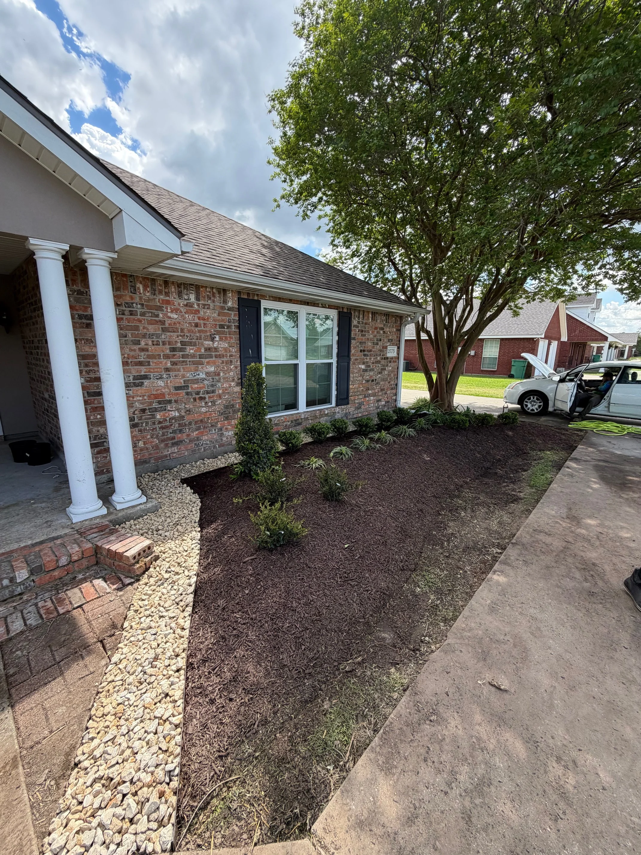 Front yard of a brick house with a large tree, a window with shutters, and a garden bed with small plants, a tall shrub, and decorative rocks under a partly cloudy sky.