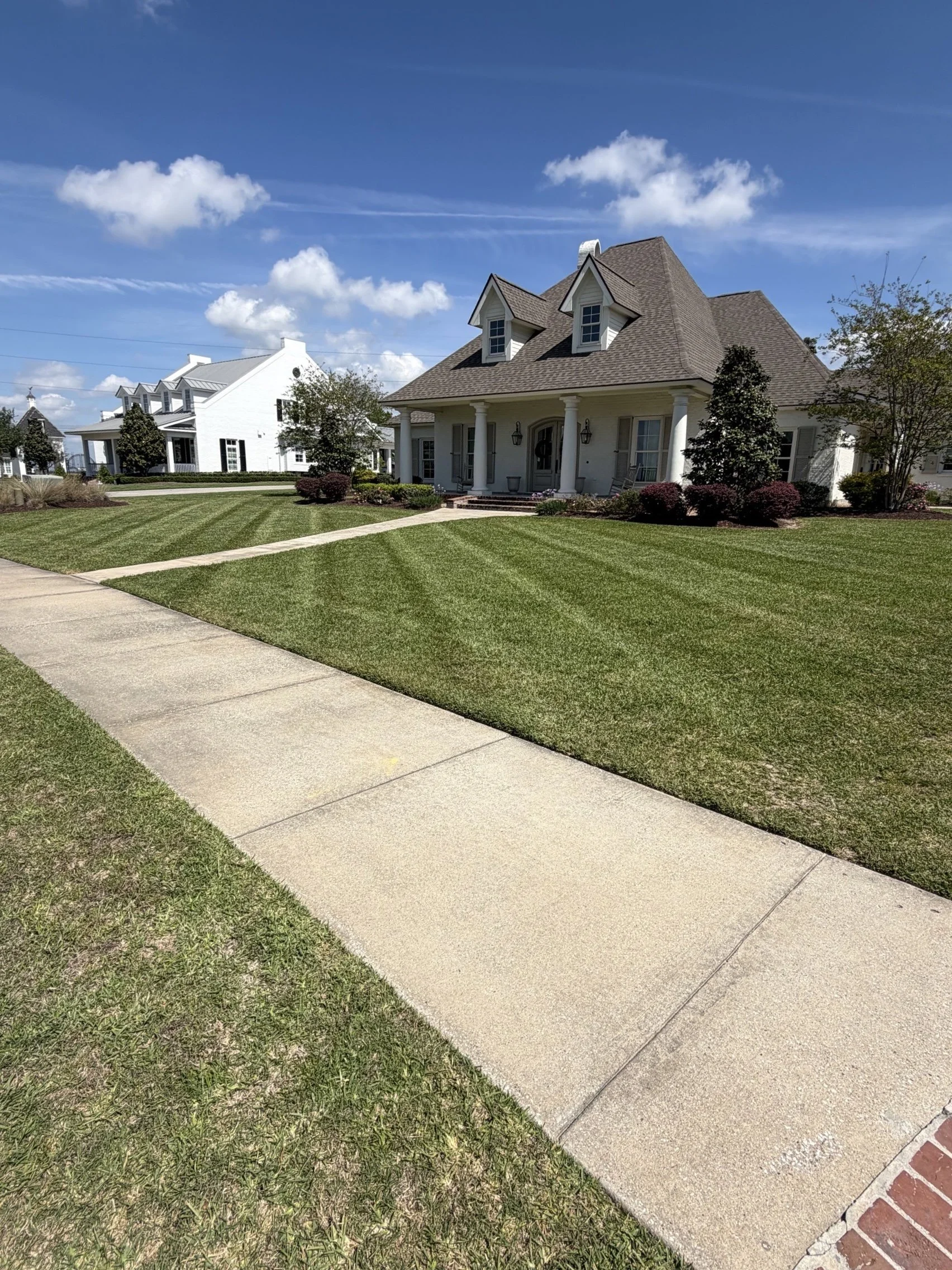 A large white house with a front porch, brown roof, and dormer windows, surrounded by a well-manicured lawn and landscaping, under a partly cloudy blue sky.