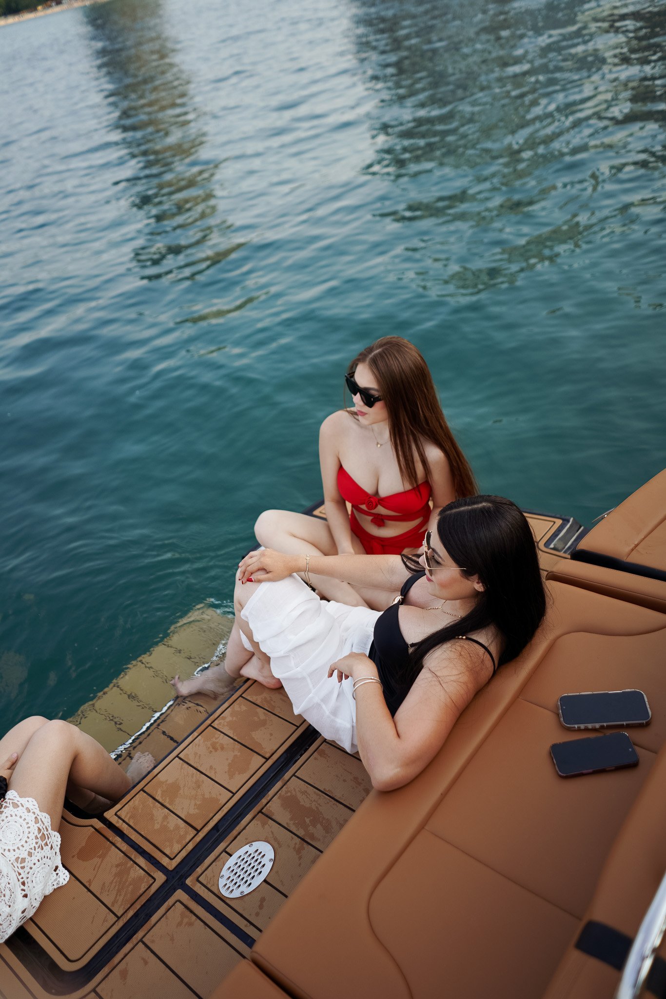 Three women sitting on a boat dock near water, one wearing a red bikini and sunglasses, another in black top and white skirt, and the third with a white lace dress, with mobile phones nearby.