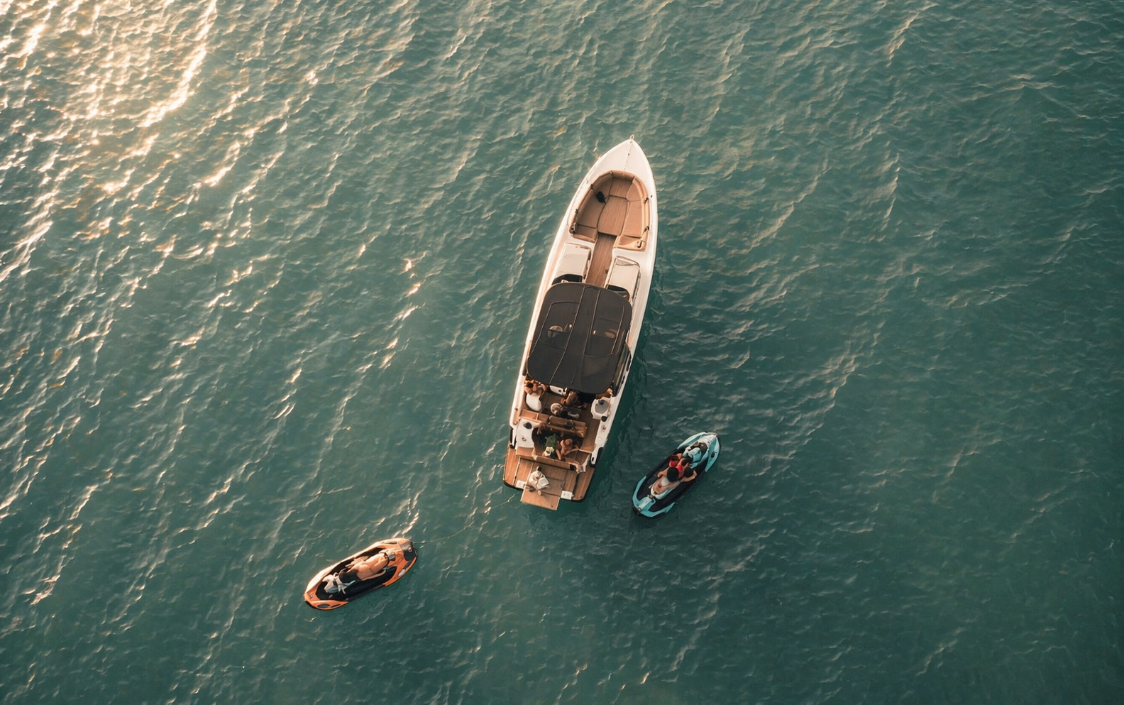 Overhead view of a white yacht with a black canopy, surrounded by two jet skis with people, on calm water during sunset.