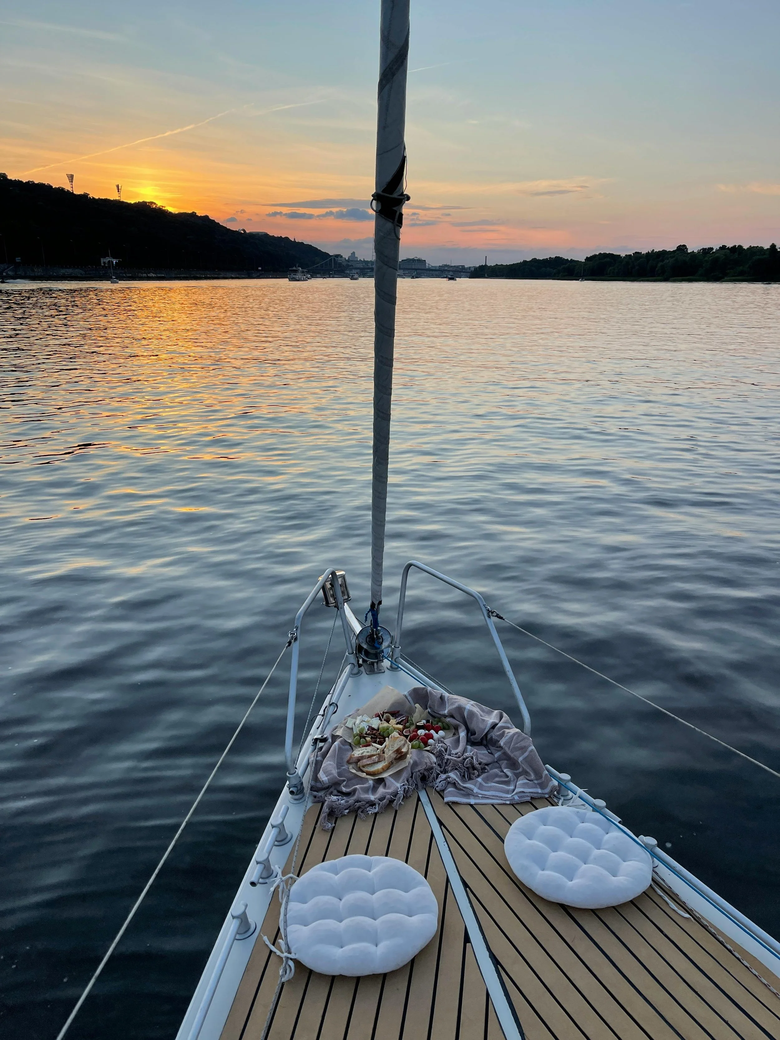View from a sailboat at sunset with a calm river, hills on either side, and multiple boats in the water. The boat's deck has cushions and a colorful snack tray with snacks and fruits.