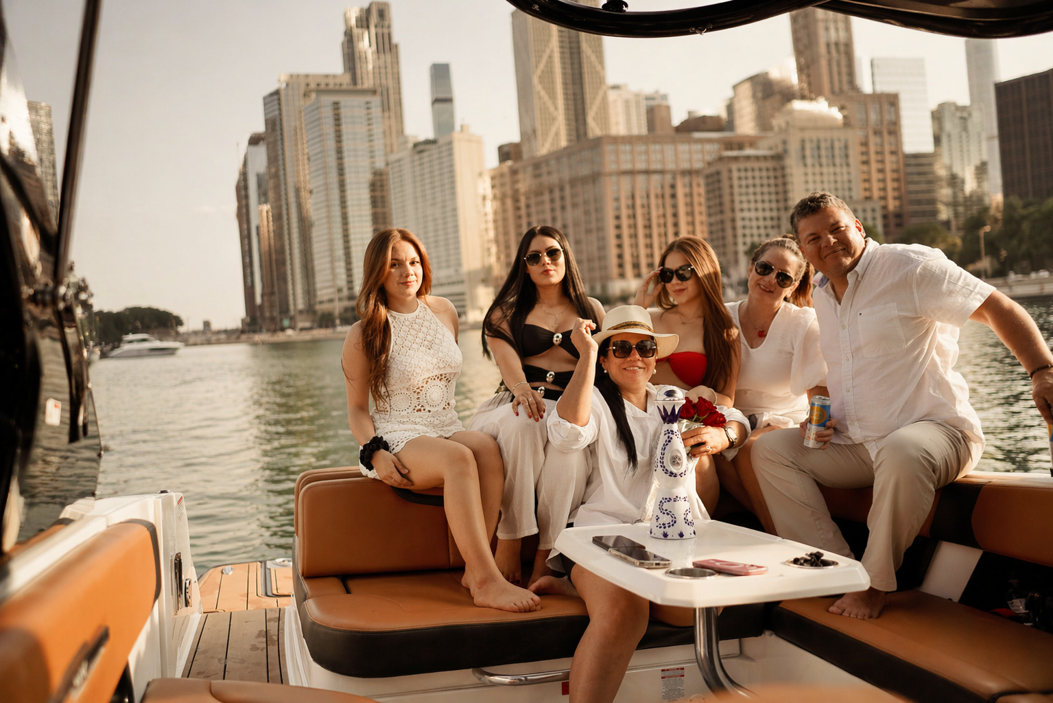 Group of six people enjoying a boat ride on a river with city skyscrapers in the background, some wearing sunglasses and summer clothing.