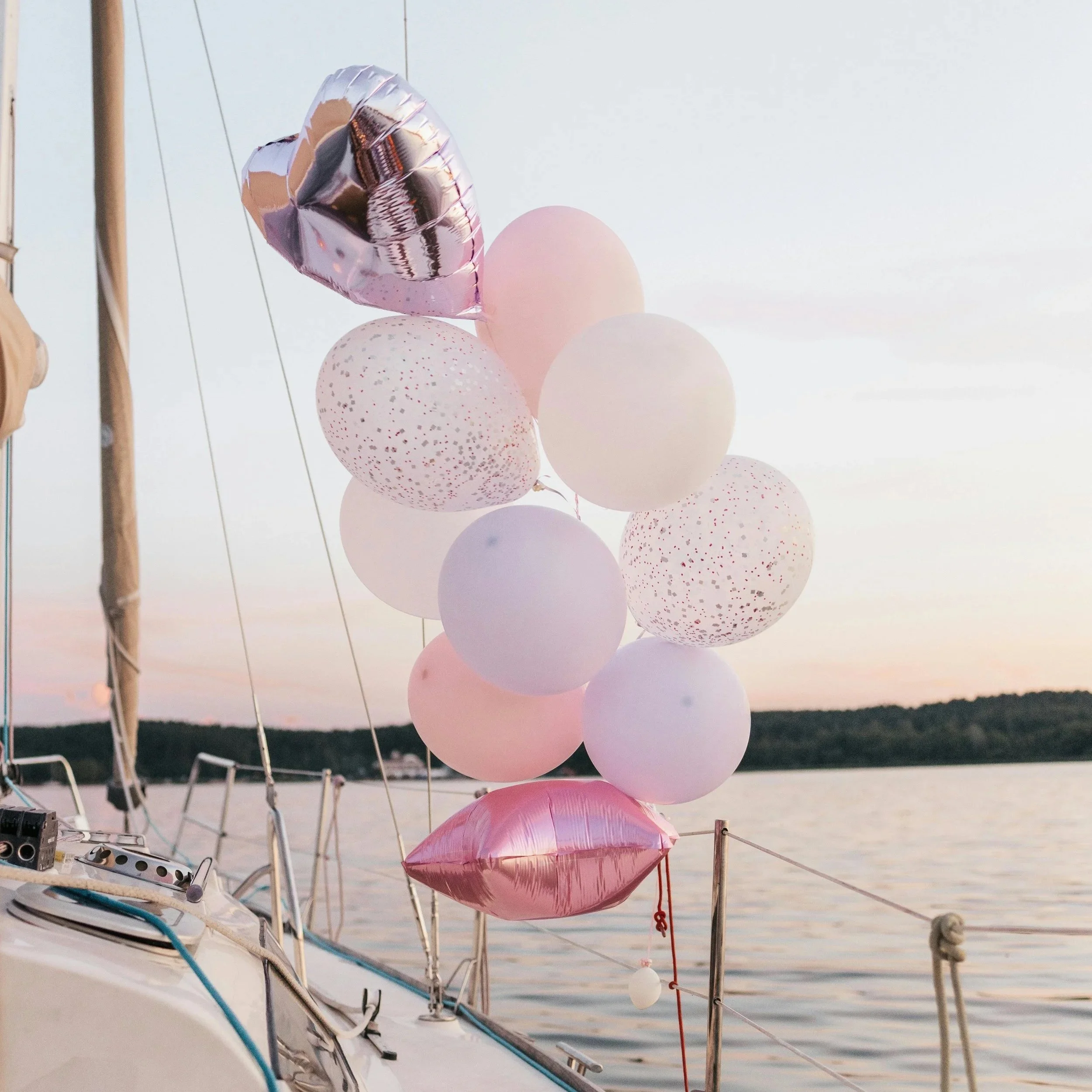 A collection of pink, white, and metallic heart-shaped balloons tied to a docked boat, with a lake and evening sky in the background.