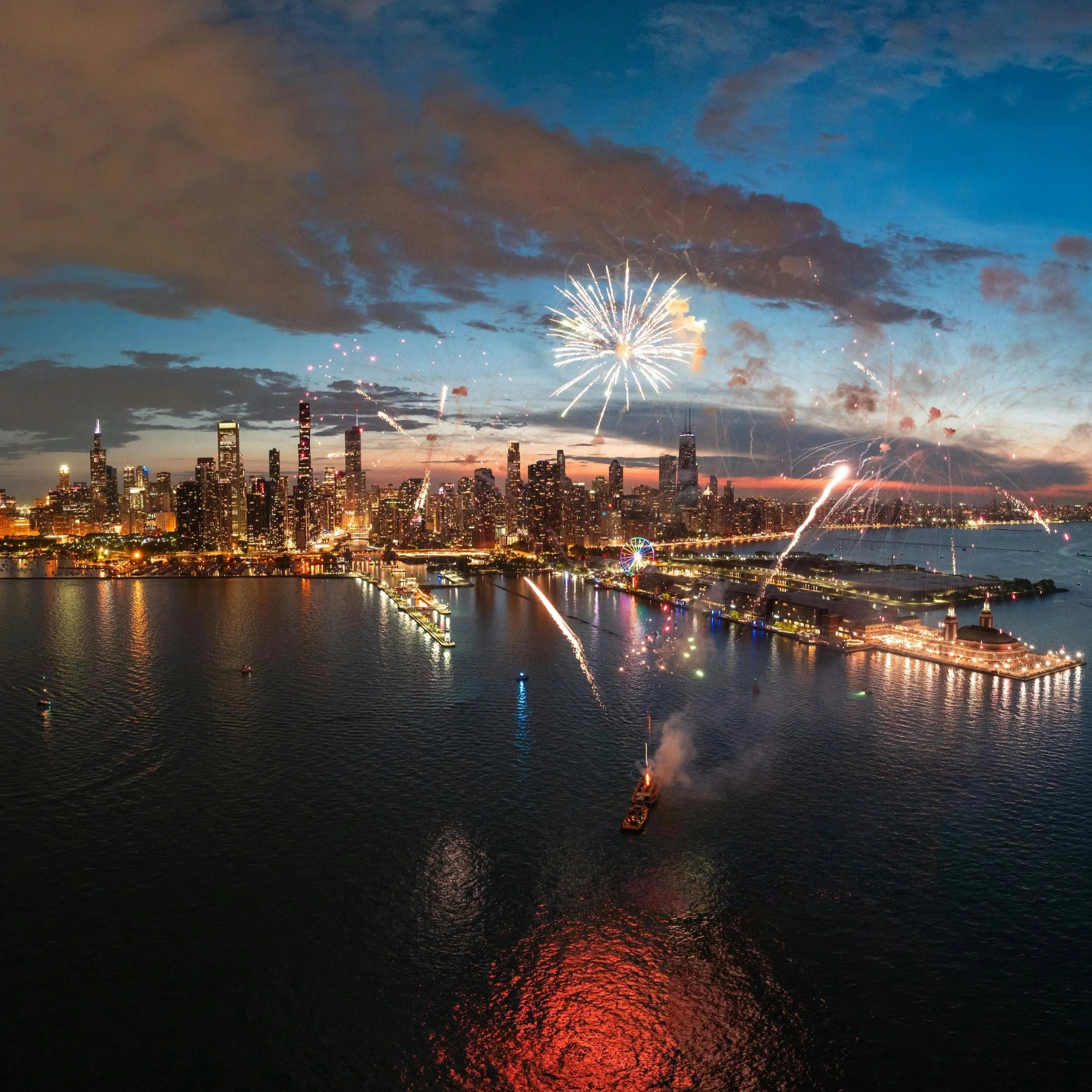Fireworks explode over a city skyline at dusk, reflecting off a river in the foreground.
