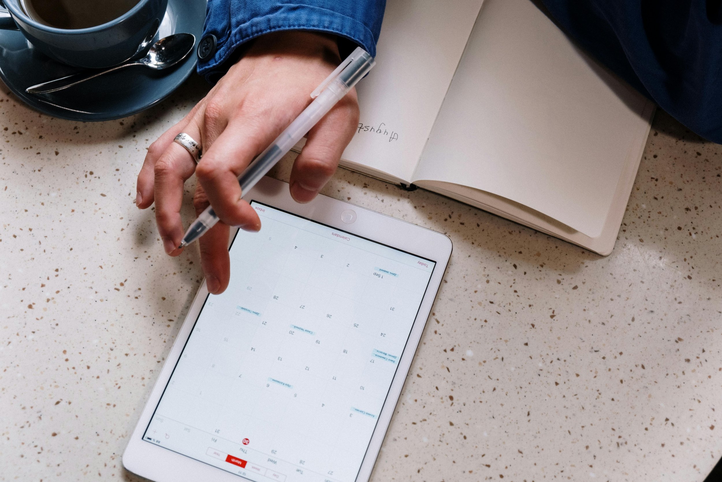 A person writing in a notebook with a pen while sitting at a speckled countertop, next to a tray with a cup of coffee and a spoon. A smartphone displaying a calendar app is also visible on the countertop.