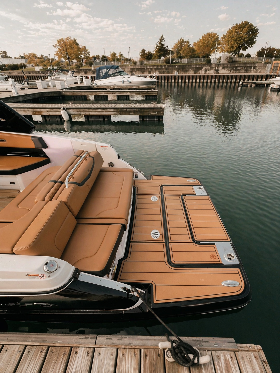 View of a marina with a brown cushioned boat seat on a boat docked near a floating wooden dock. Several boats are moored in the background, including a white motorboat and other smaller boats, with a calm water surface and trees with fall foliage in 