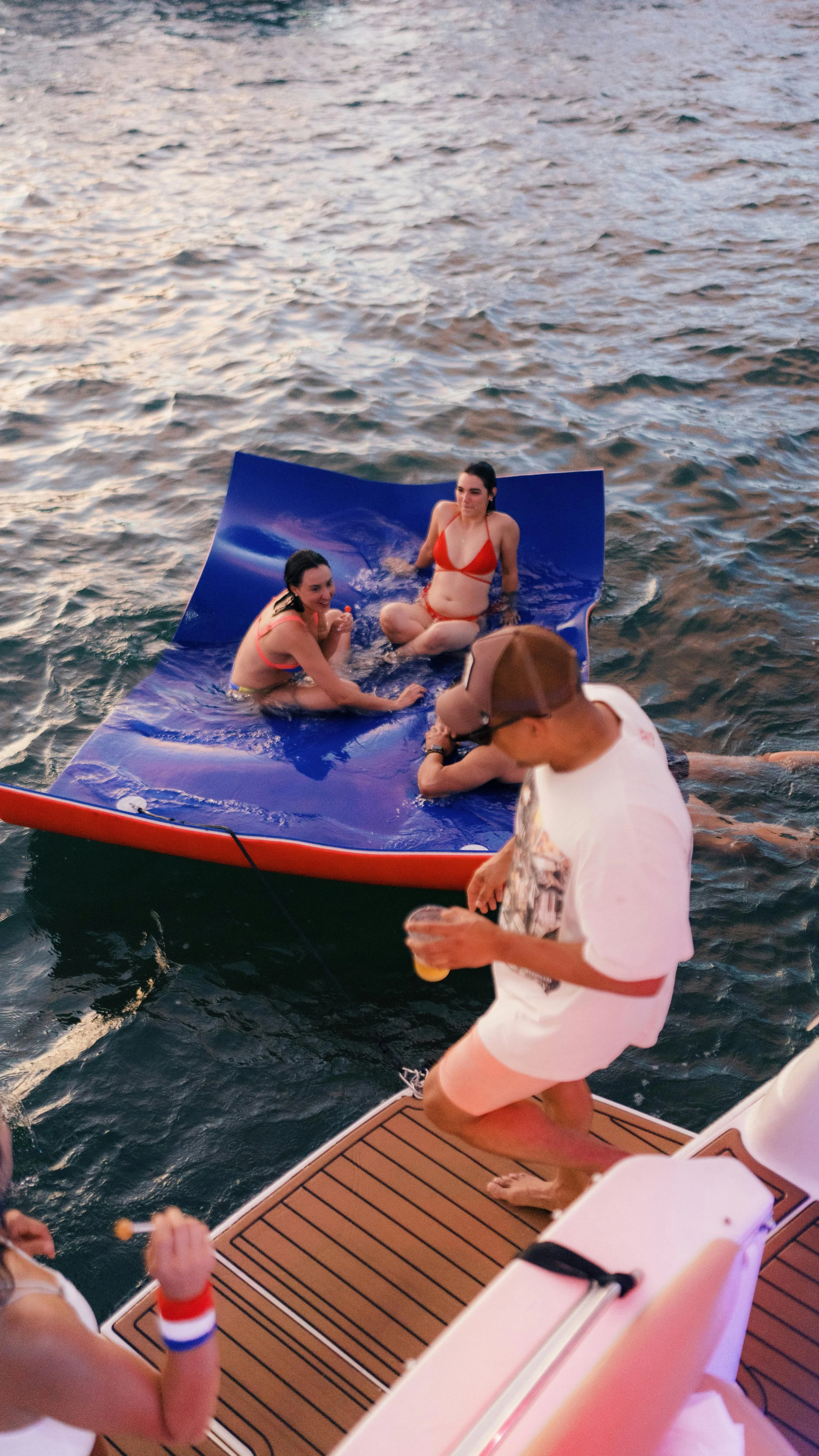 Two women in swimsuits sitting on a floating mat in the water, with a person standing nearby holding a drink and wearing a cap, on a boat dock at dusk.