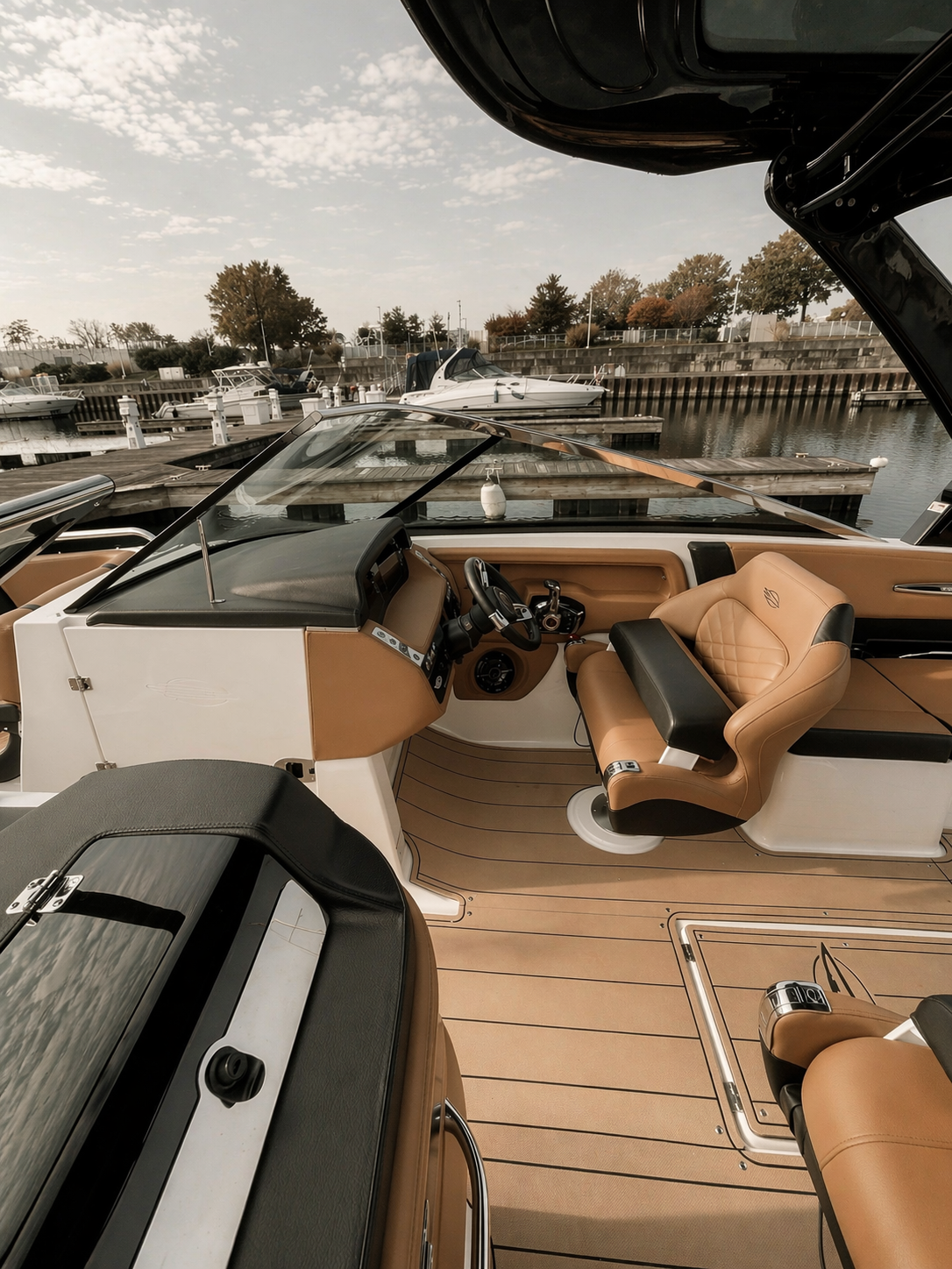 Interior view of a luxury boat with beige and black seating, steering wheel, and dashboard, docked in a marina with other boats and trees in the background.