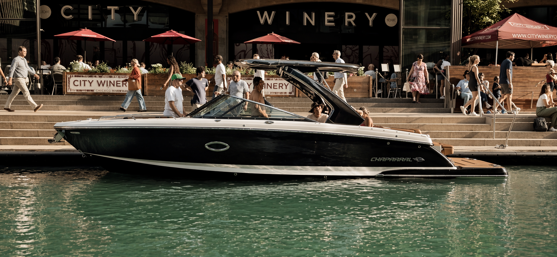 A black and white motor yacht named Chaparral docked along a city waterfront with people walking and sitting on steps outside a restaurant labeled City Winery.