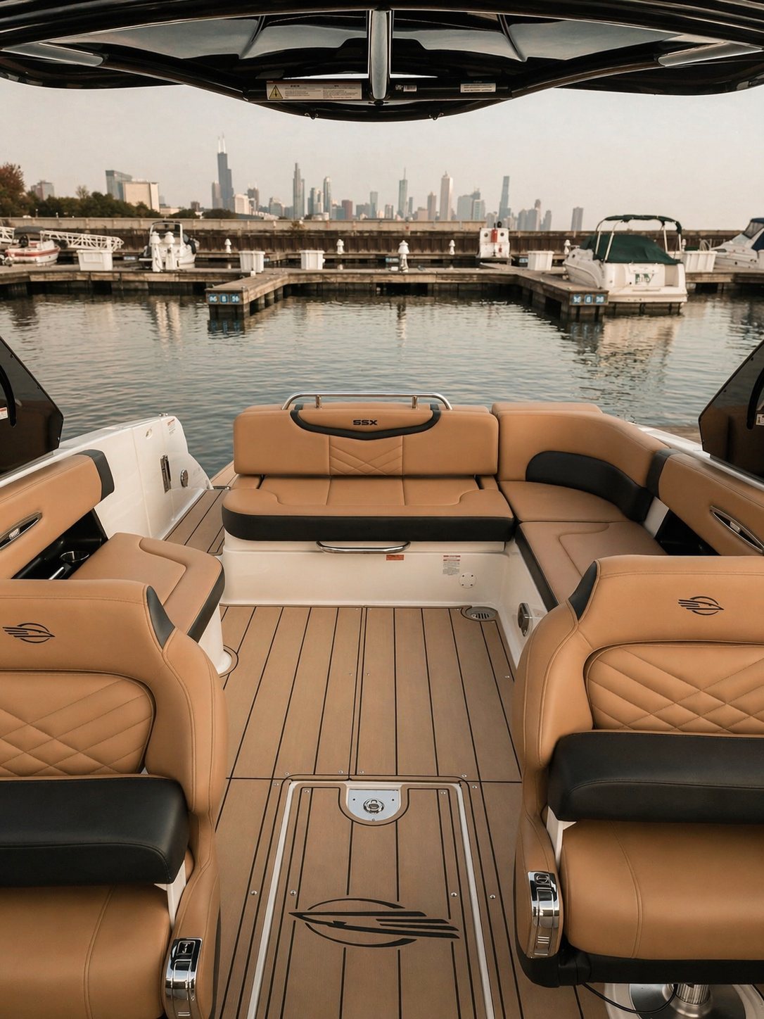 View from the deck of a luxury boat looking out over a marina with Chicago skyline in the background, including the Willis Tower and other high-rise buildings. The boat's interior features beige and black cushioned seats with a wood-patterned floor.