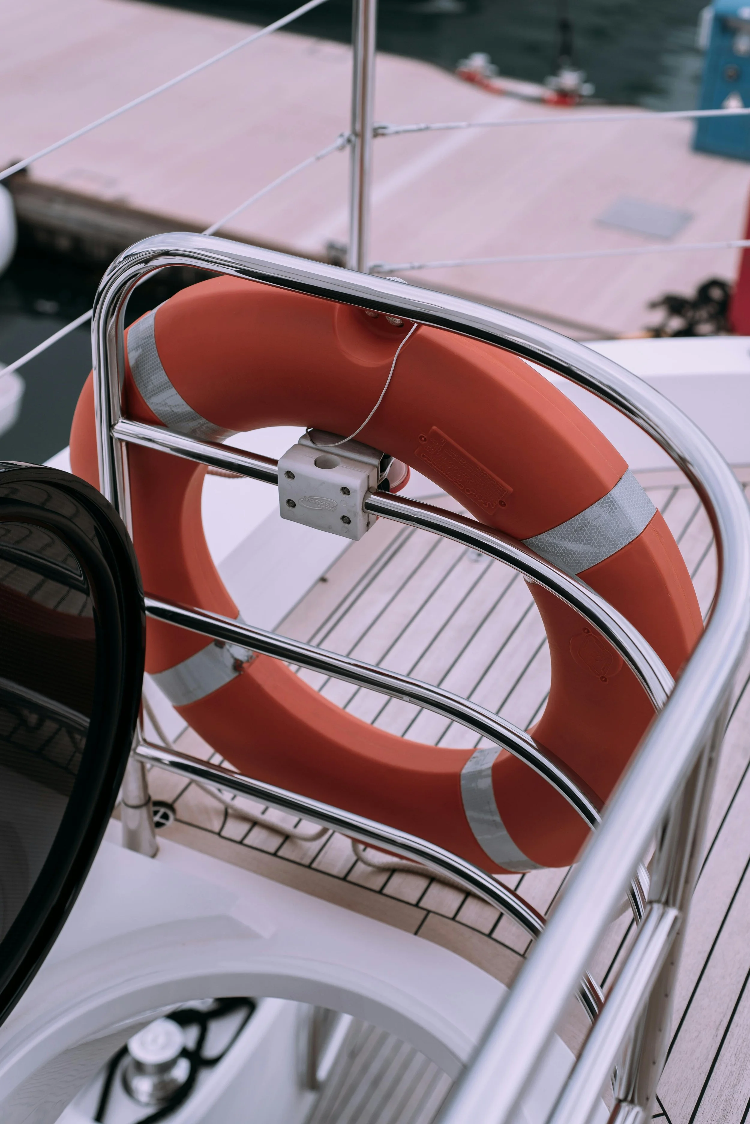 Close-up of an orange lifebuoy with reflective strips, secured to a boat's metal railing, with a black wheel and parts of the boat deck visible in the foreground, and water in the background.