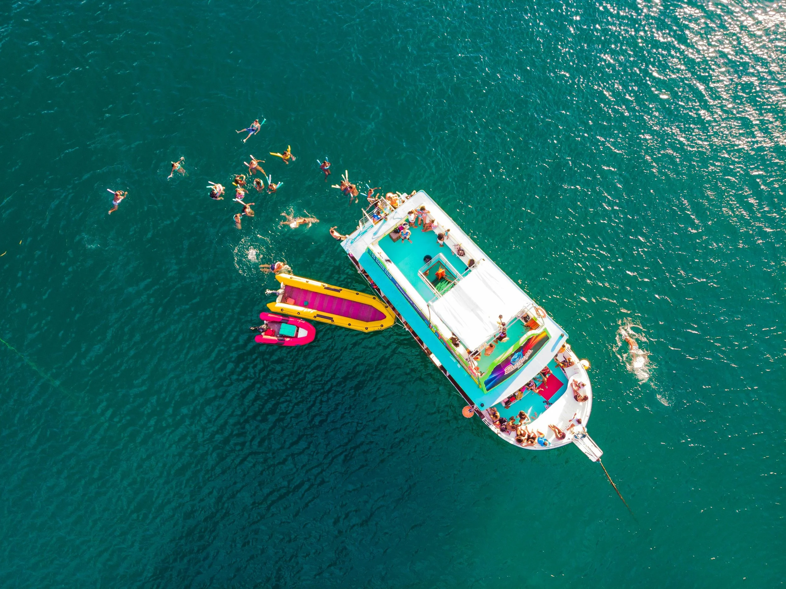 An aerial view of a boat with colorful slides and a pool, carrying multiple people, floating on a large body of water. Several individuals are swimming nearby, enjoying a sunny day.