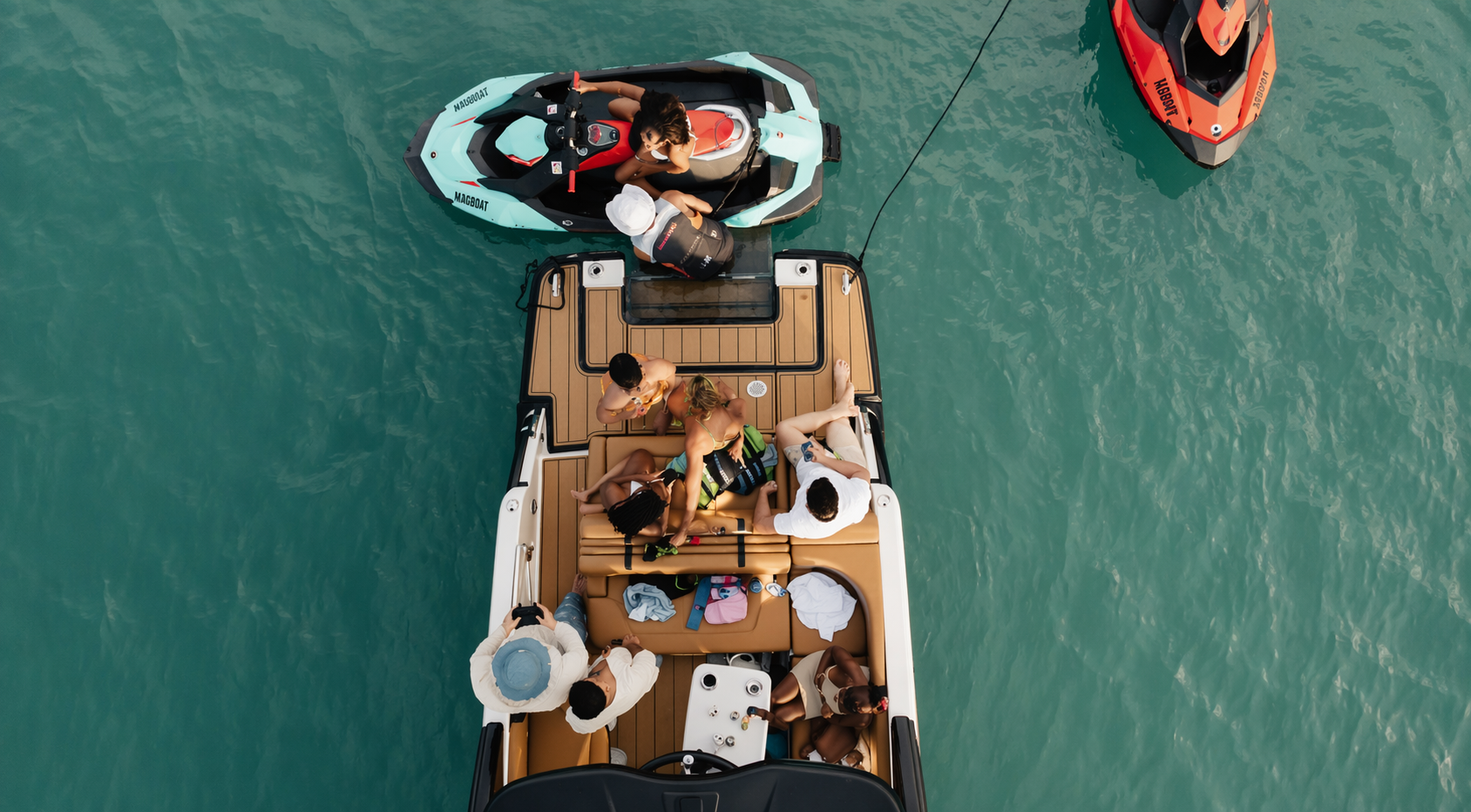 Overhead view of a boat with seven people on board, some sitting and some standing. Two people are on a jet ski connected to the boat. The boat is on turquoise water, and there is another boat visible in the top right corner.