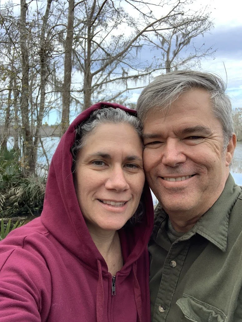 A smiling man and woman taking a selfie outdoors near a lake or river with trees in the background, cloudy sky, and the woman wearing a maroon hoodie.