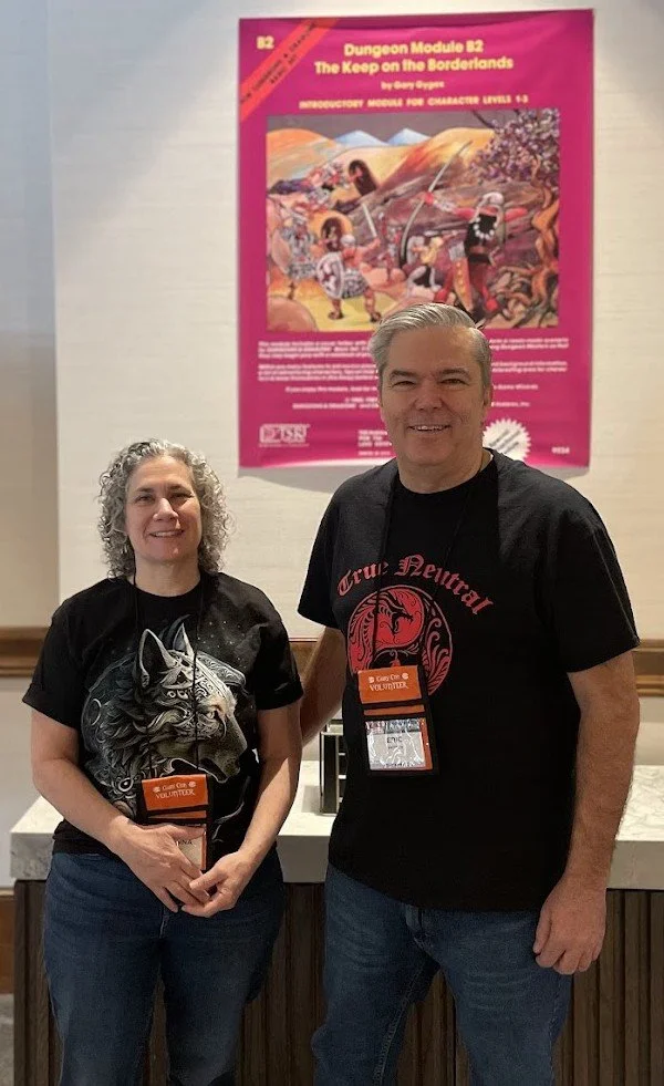 Two people standing in front of a colorful poster for the Dungeon Module B2 titled 'The Keep on the Borderlands.' The woman on the left has curly gray hair and is wearing a black T-shirt with a wolf design, and the man on the right has gray hair and is wearing a black T-shirt with red text and graphics. Both are wearing volunteer name badges.
