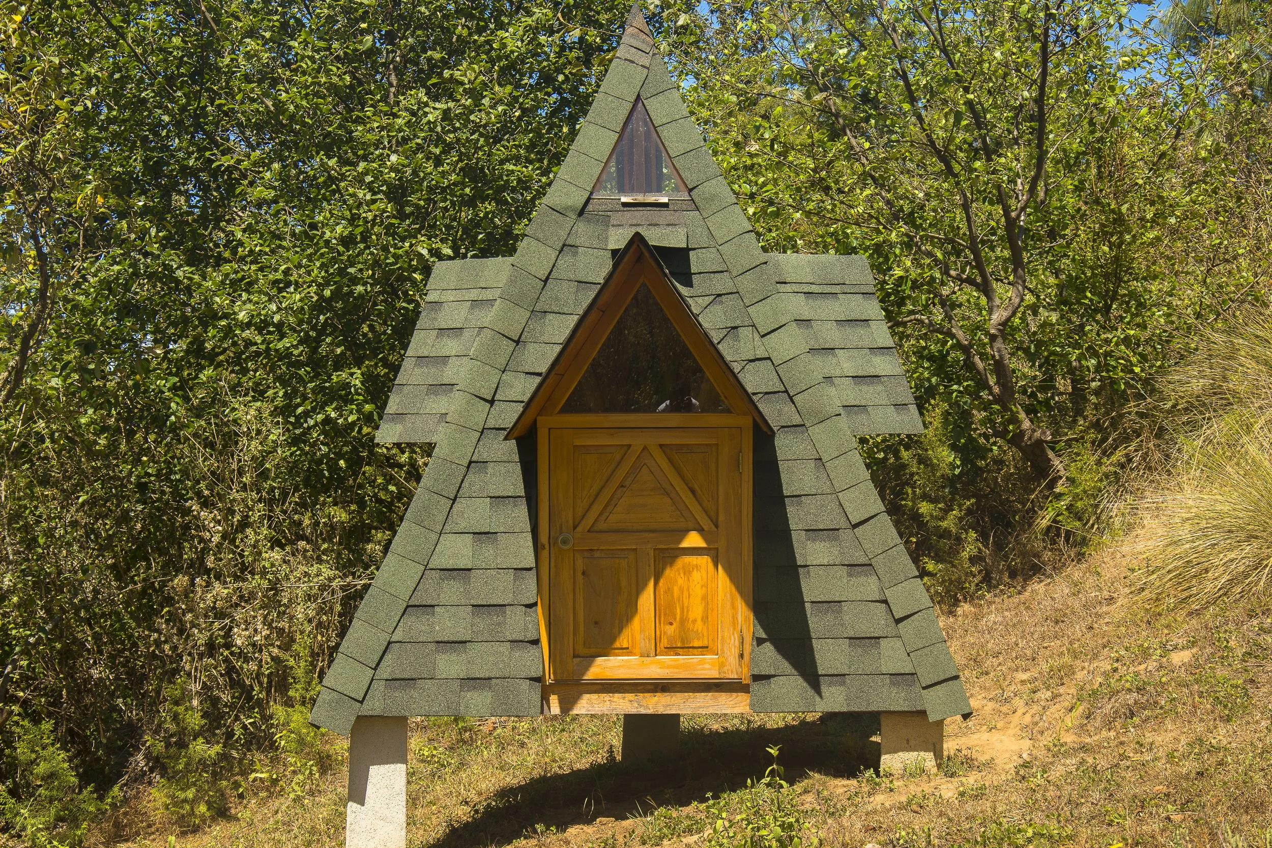Casita de madera con techo a dos aguas y ventanas en forma de triángulo, rodeada de árboles verdes.