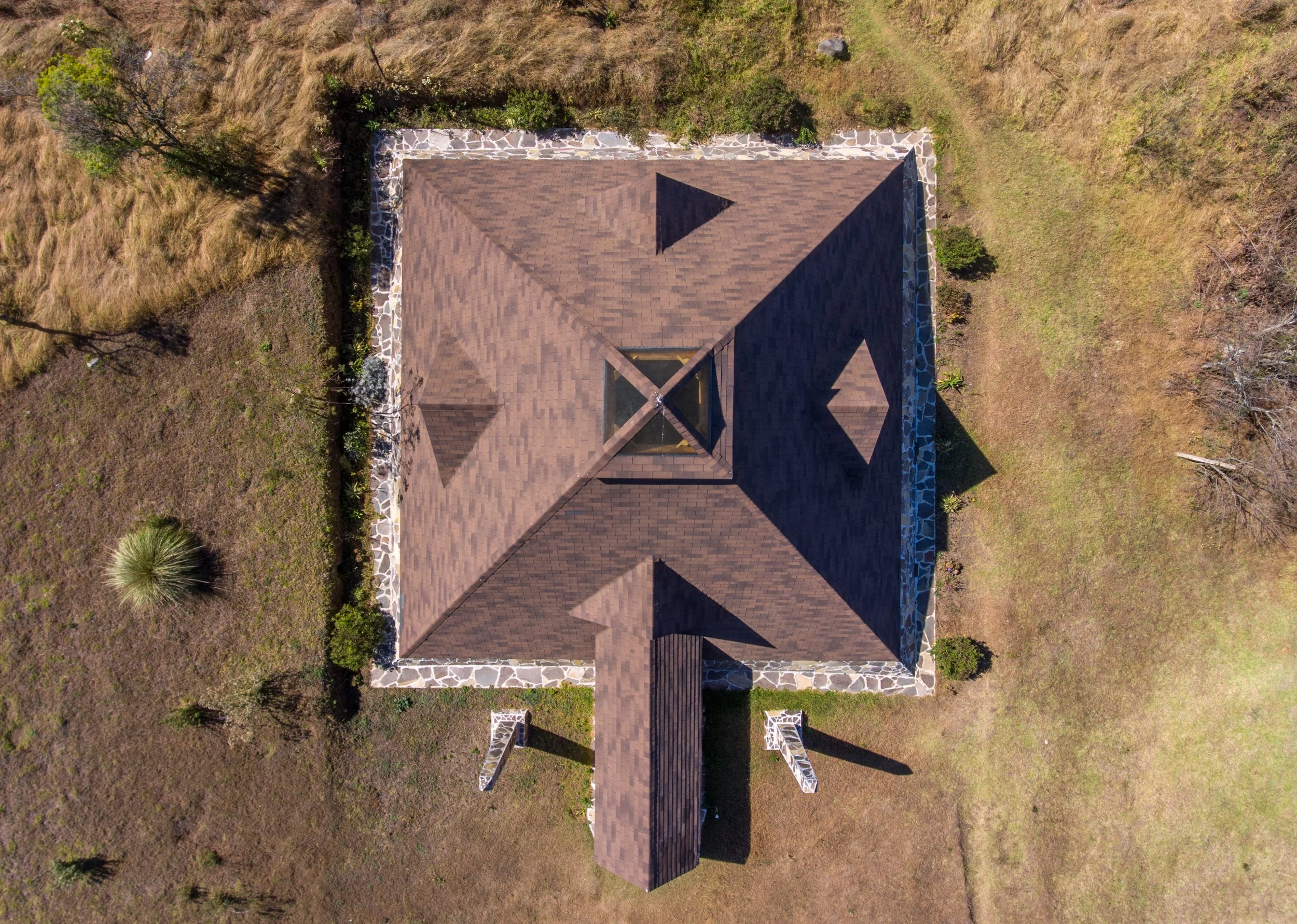 Vista aérea de una vivienda con techo de tejas marrones, rodeada de un marco de piedra y terrenos con césped y árboles alrededor.