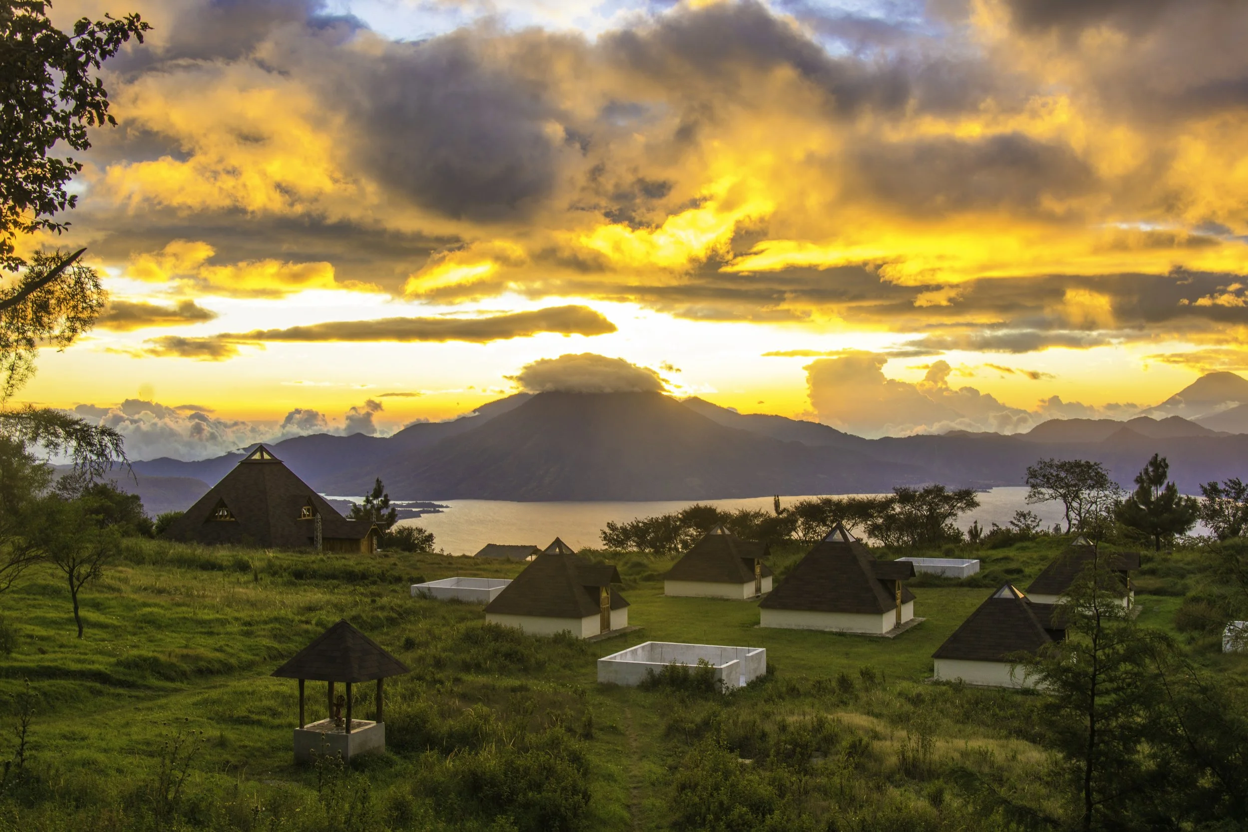 Paisaje al atardecer con nubes naranjas, montañas y un lago en el fondo. Casas con techos cónicos en primer plano, rodeadas de vegetación.