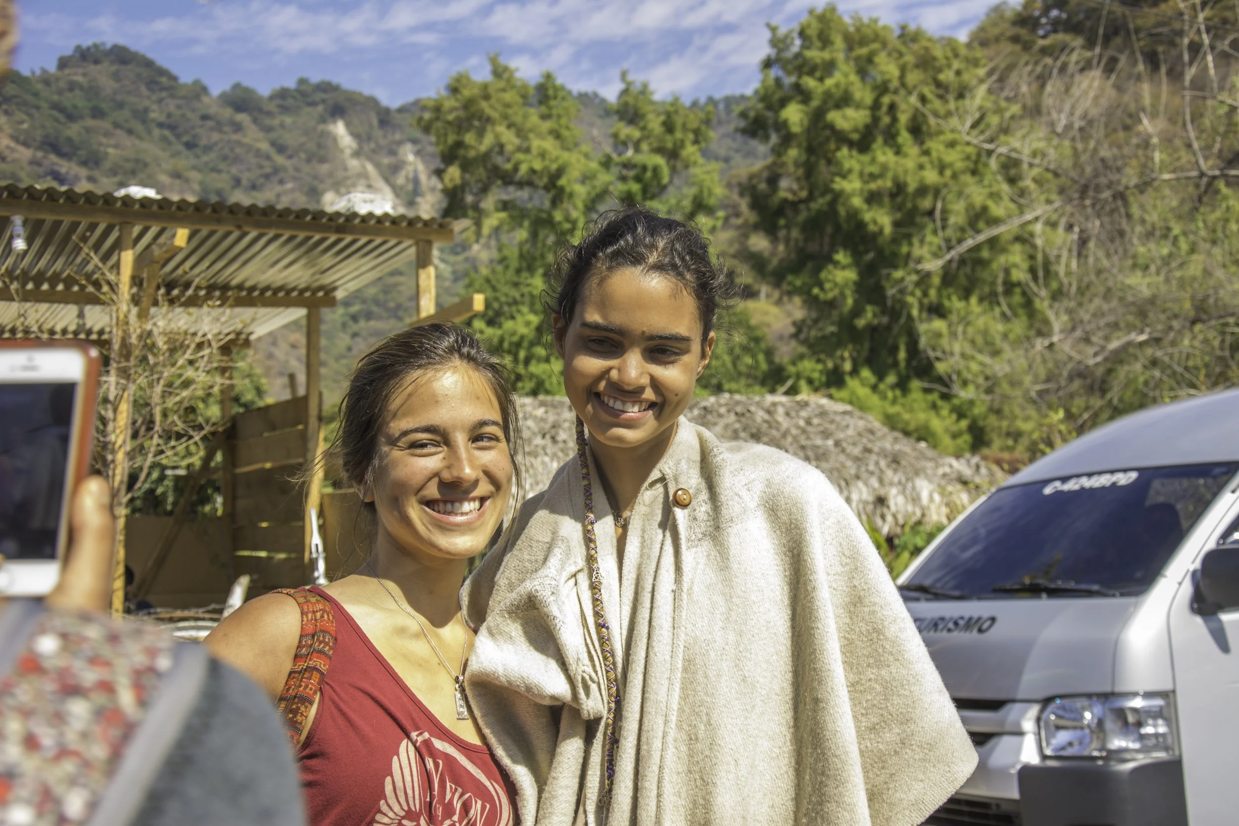 Dos mujeres sonriendo en un paisaje de montañas y árboles, con un vehículo plateado detrás y una estructura de madera, mientras alguien toma una foto con un teléfono móvil.