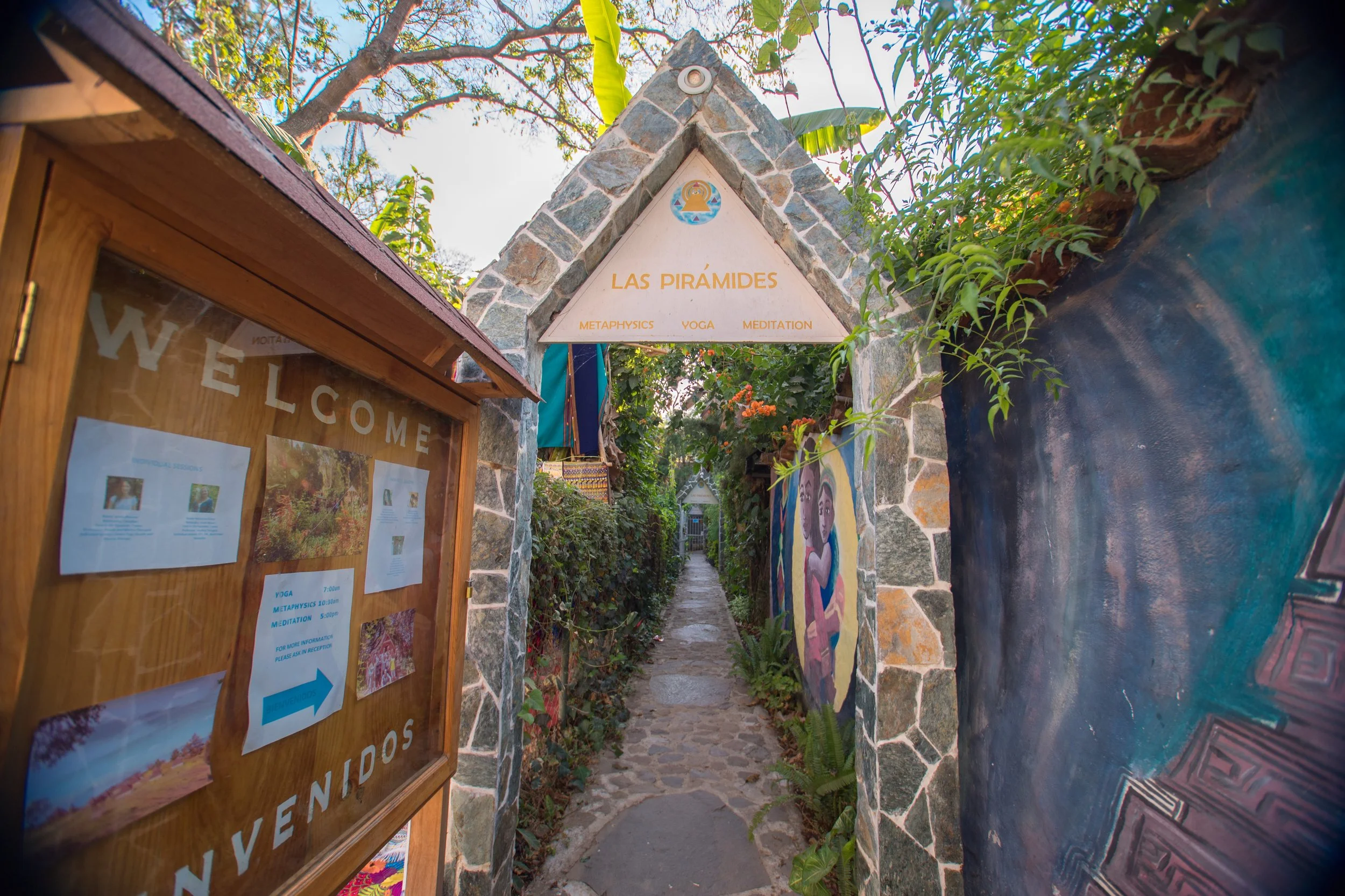Entrada a un espacio dedicado a las pirámides con un arco de piedra y árboles alrededor, cartel informativo de madera con horarios y actividades como yoga, meditación y metafísica, y una pintura de una mujer en la pared.