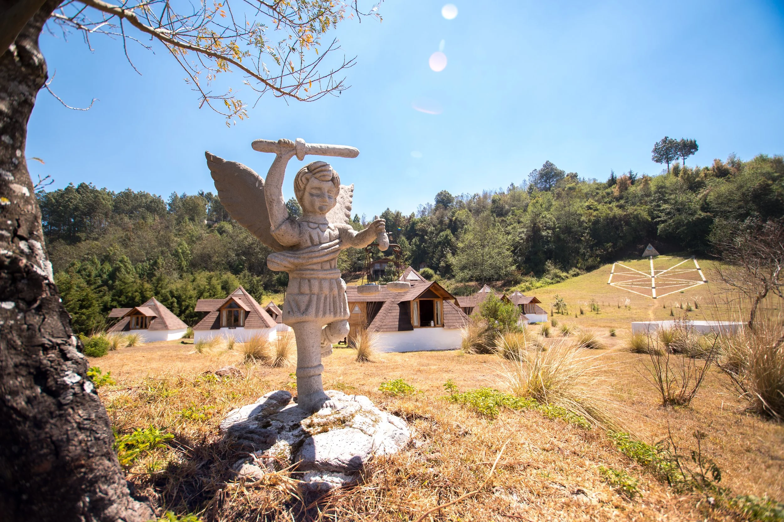 Escultura de un hada con alas, sosteniendo una especie de espada o lanza, en un entorno natural con árboles, casas de forma cónica, y un cielo despejado con el sol brillante.
