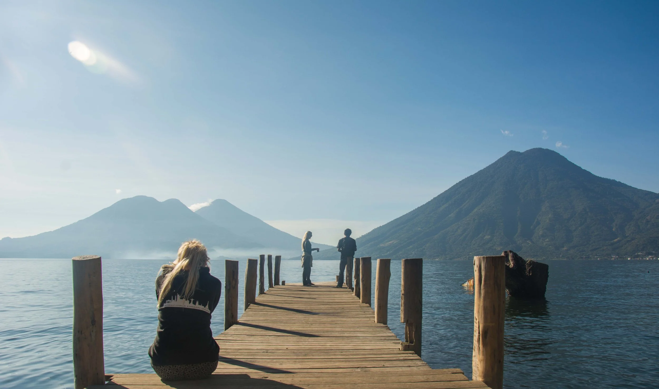 Mujer sentada en un muelle de madera frente a un lago, con dos personas de pie hablando, y montañas en el fondo bajo un cielo despejado.