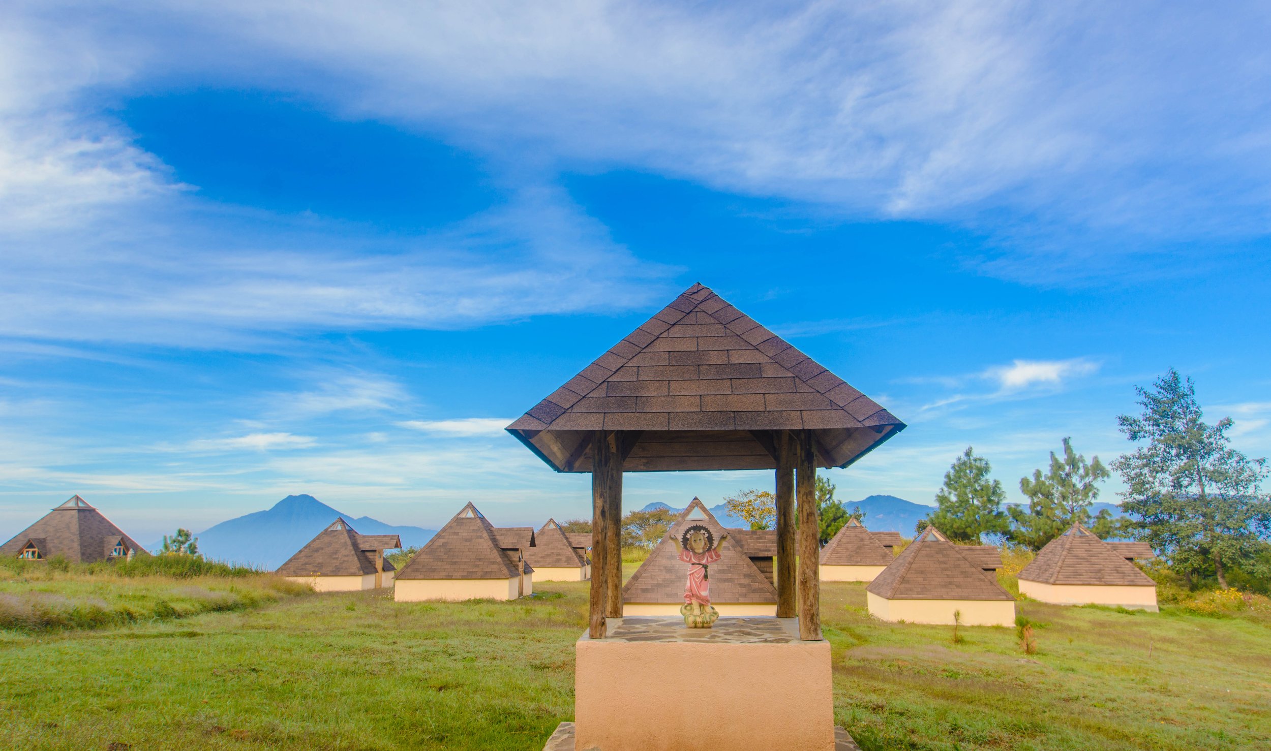 Estructura de madera con techo en forma de pirámide en un campo con varias casas de techo de tejas, árboles y montañas al fondo, cielo azul con nubes.