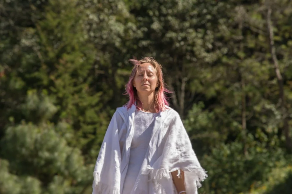 Mujer con cabello rubio y rubio con puntas rosa, usando una blusa blanca, caminando por un bosque con árboles verdes.