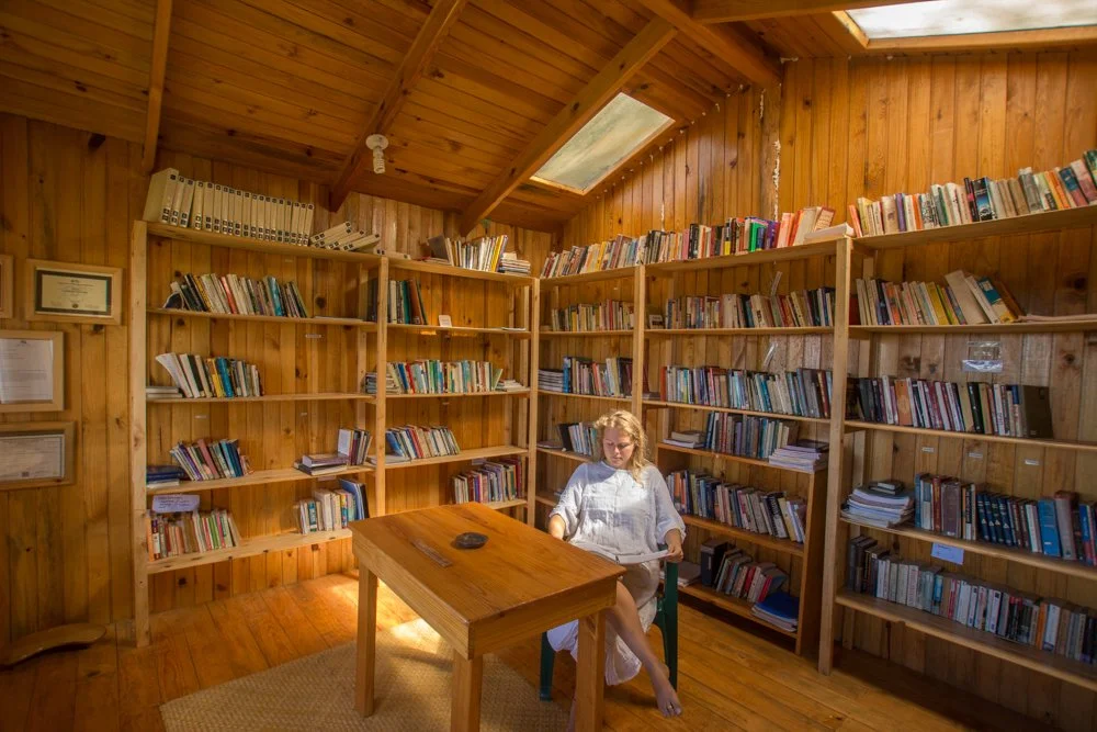 Biblioteca de madera con estanterías llenas de libros, una mujer sentada en una silla leyendo en una mesa en el centro, techo de madera con ventanas y luz natural.
