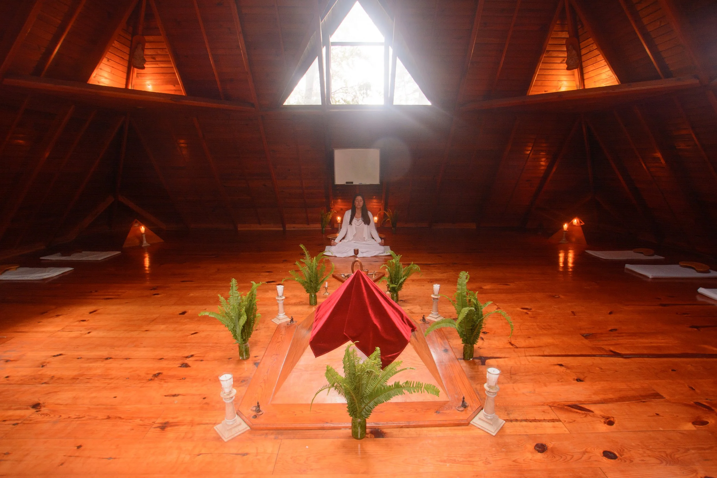 Una mujer sentada en posición de meditación en un espacio de madera con paredes inclinadas, rodeada de velas y plantas, con luz natural entrando por una ventana en el techo.