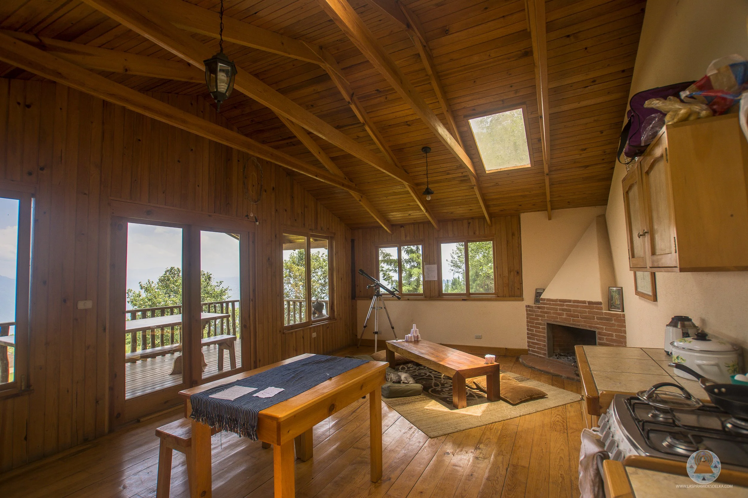Interior de una cabaña de madera con paredes y techos de madera, ventanas grandes y una abertura en el techo con vista al cielo. Hay muebles de madera, una mesa y un banco, y un telescopio junto a la ventana.