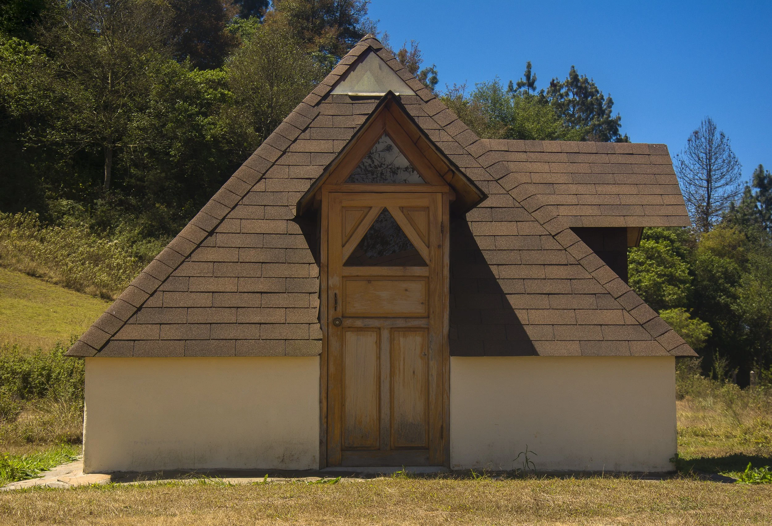 Casa con techo a dos aguas y una pequeña ventana triangular en la parte superior, rodeada de árboles y vegetación.