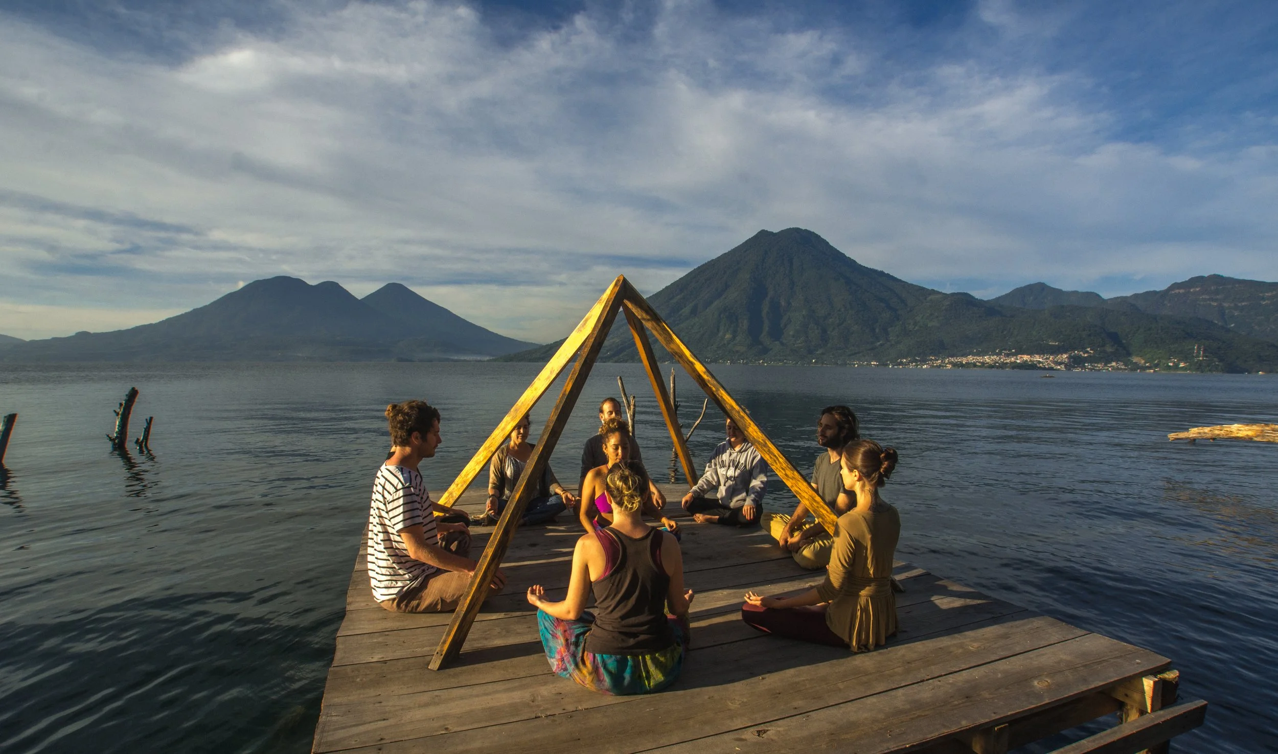 Grupo de personas sentadas en círculo en un muelle de madera en un lago cercano a un volcán, participando en una actividad de meditación o yoga, con montañas y nubes en el cielo al fondo.