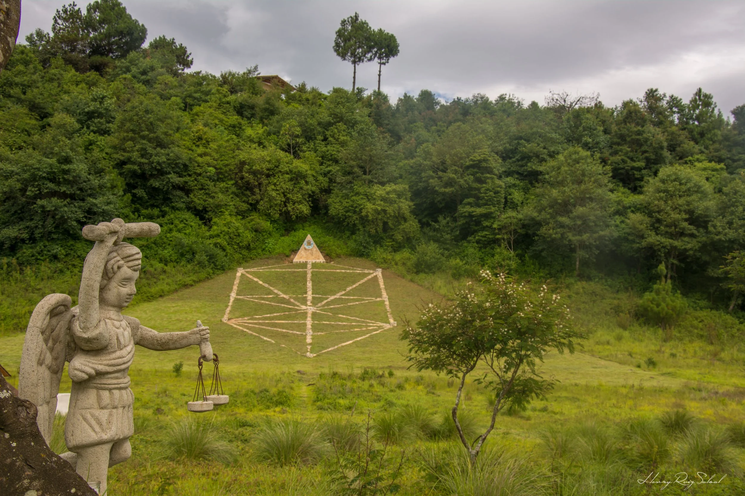 Escultura de piedra de una figura indígena con balanza en mano en primer plano, un área con césped y árboles, una pirámide de piedra decorada con motivos azules, y una colina cubierta de árboles verdes en un día nublado.
