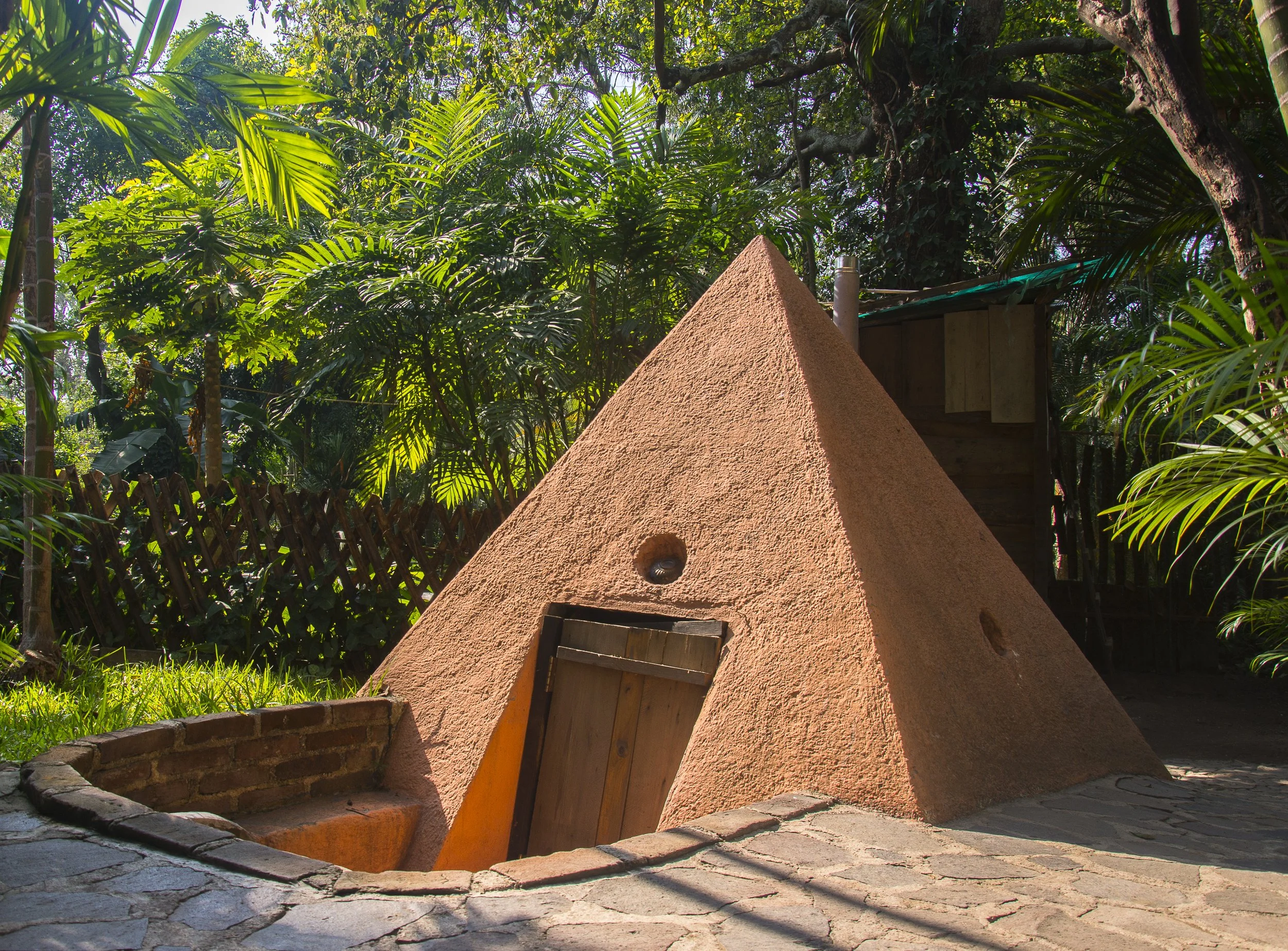 Estructura de barro en forma de pirámide en un jardín rodeado de plantas verdes.