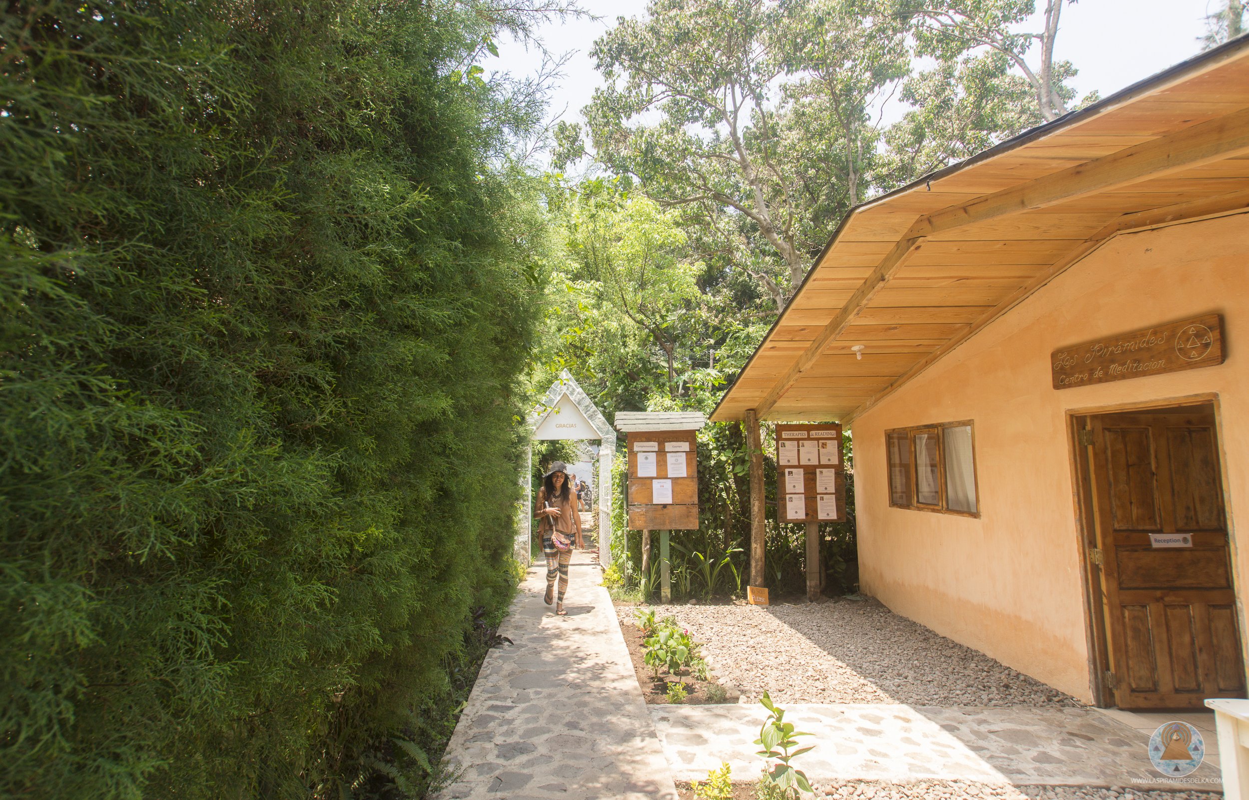 Camino de piedra junto a un muro de arbustos verdes a la izquierda, dirigidamente a un edificio en color naranja con ventanas de madera y un cartel que dice 'Los Pirámides Centro de Meditación'. Personas caminando por el sendero, rodeadas de árboles.