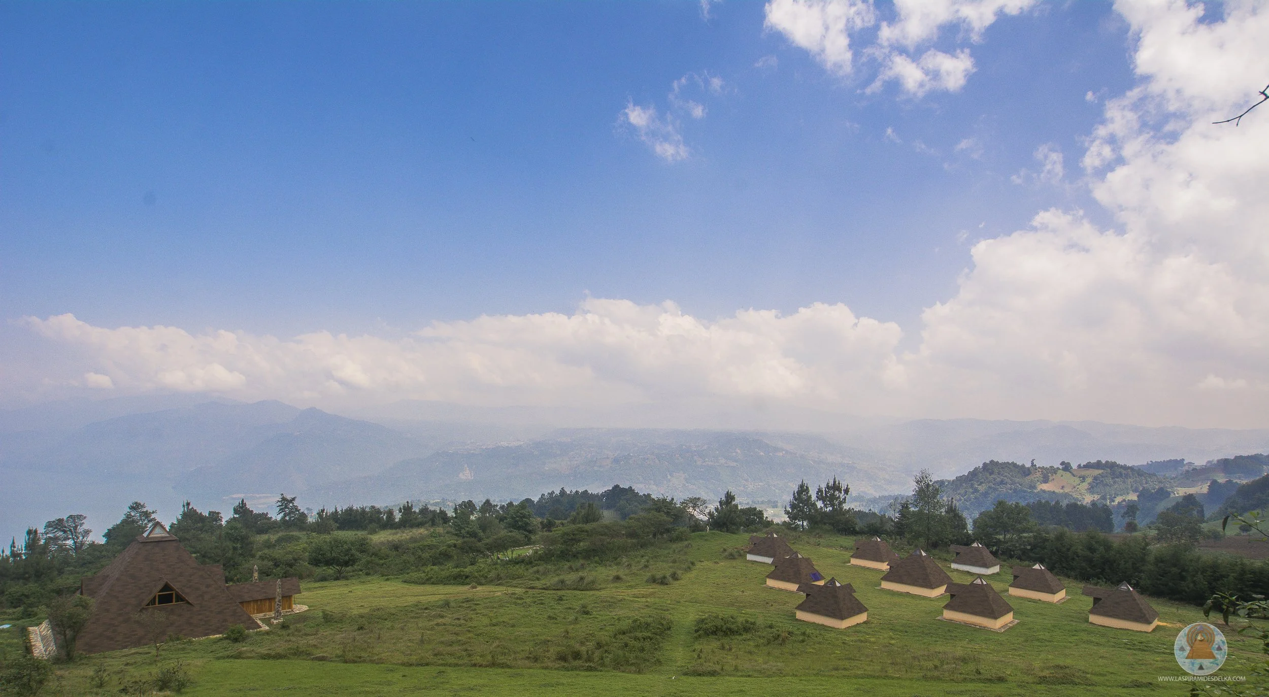 Paisaje rural con casas de techos de paja y vegetación, montañas y cielo con nubes blancas y un cielo azul