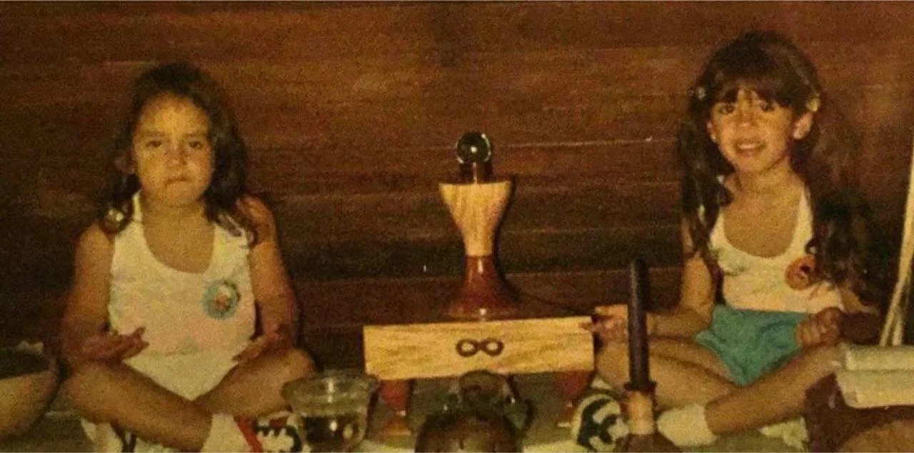 Dos niñas con cabello rizado y oscuro sentadas en una mesa con juguetes y una botella, frente a un fondo de madera.