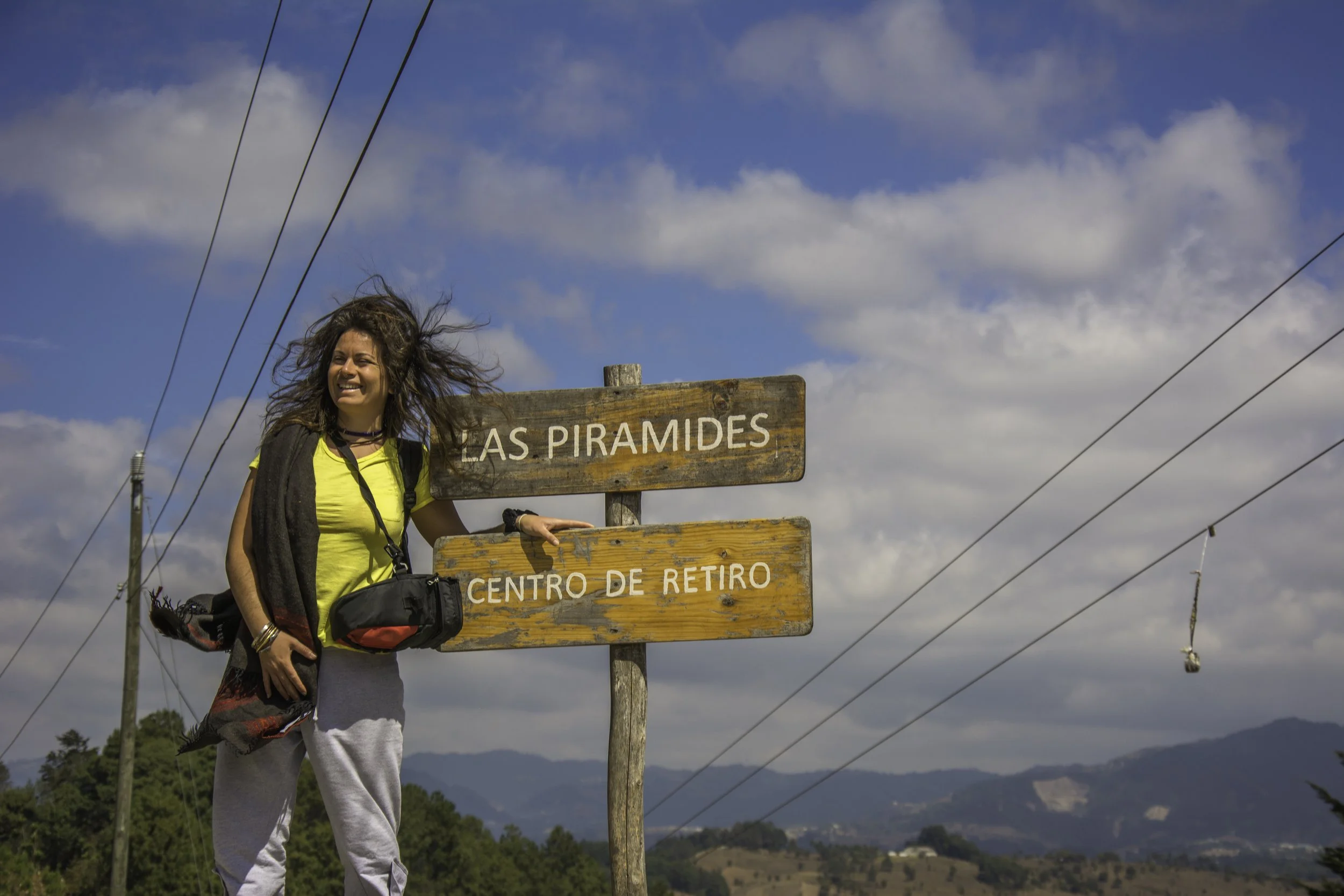 Mujer con mochila en un lugar con señal que dice "Las Pirámides" y "Centro de Retiro" bajo un cielo azul con nubes y cables eléctricos en el fondo.