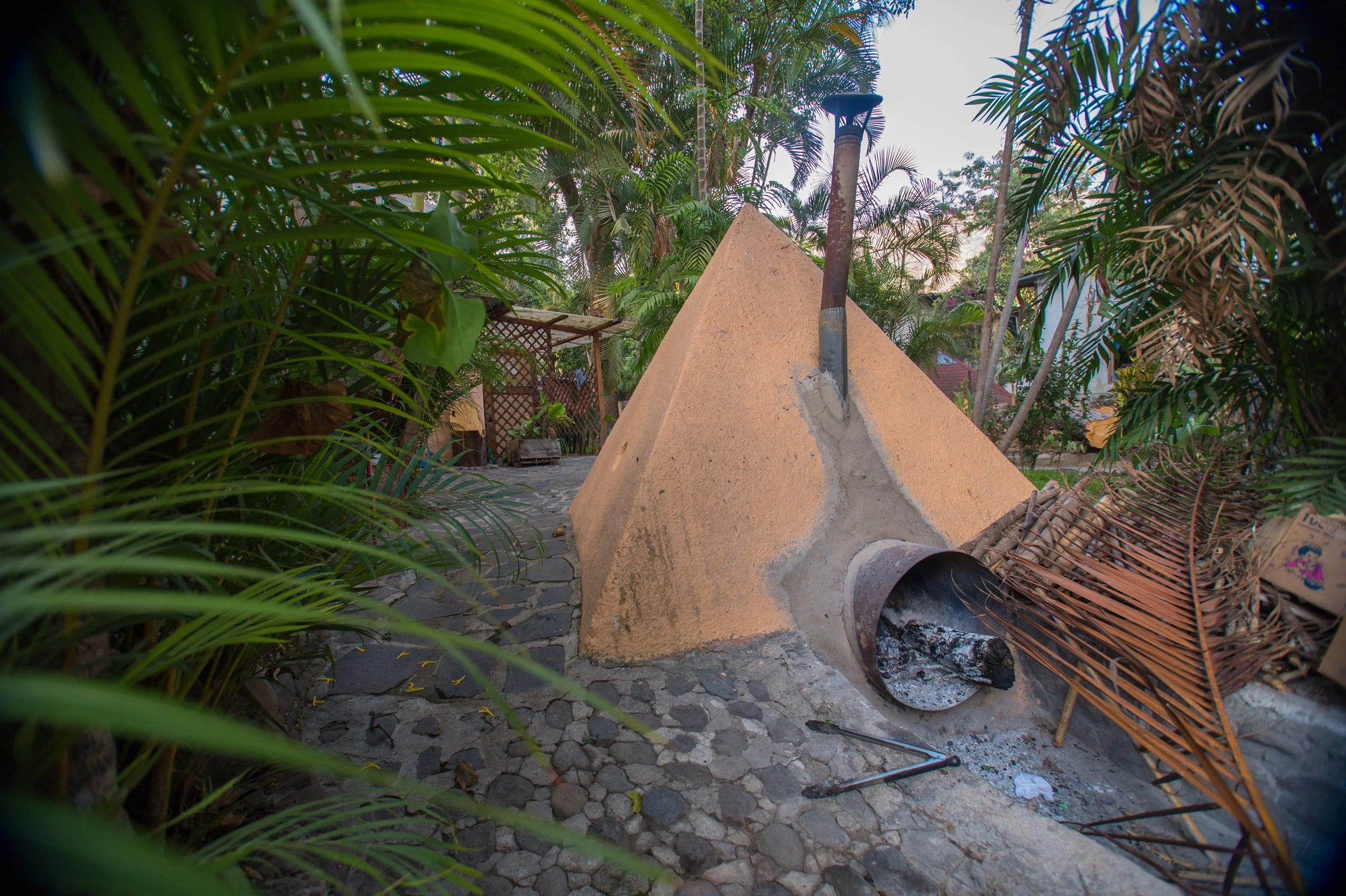 Una estructura de chimenea de barro con un largo tubo de metal en la parte superior, rodeada de plantas verdes y palmas, en un ambiente exterior natural con caminos de piedra.