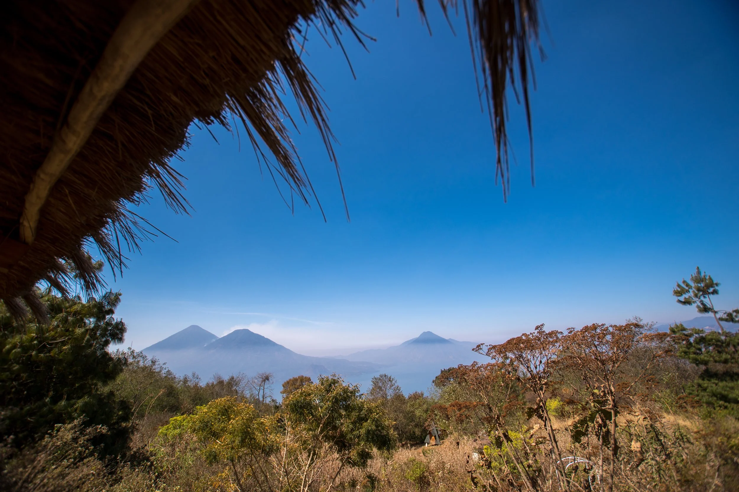 Vista panorámica de un paisaje montañoso con volcanes en el fondo, rodeado de árboles y vegetación, con cielo despejado.