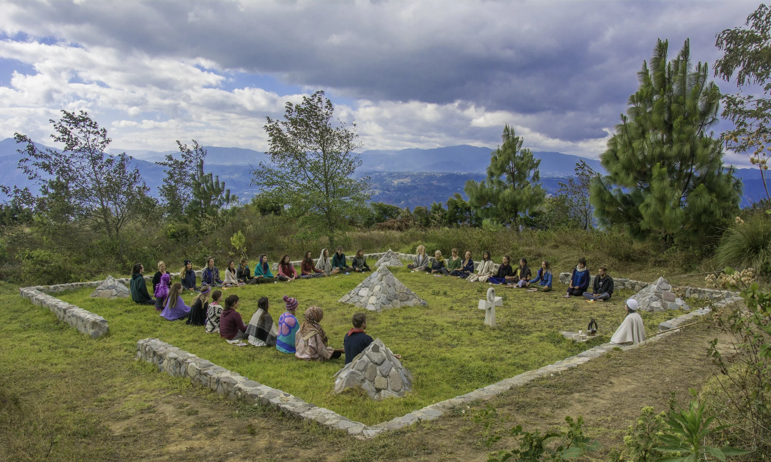 Un grupo de personas participa en una ceremonia ceremonial al aire libre en un espacio rodeado de naturaleza con árboles y montañas al fondo, en medio una estructura de piedra en forma de cruz y otro de estructuras en forma de pirámide. El cielo presenta nubes y algunos rayos de sol.